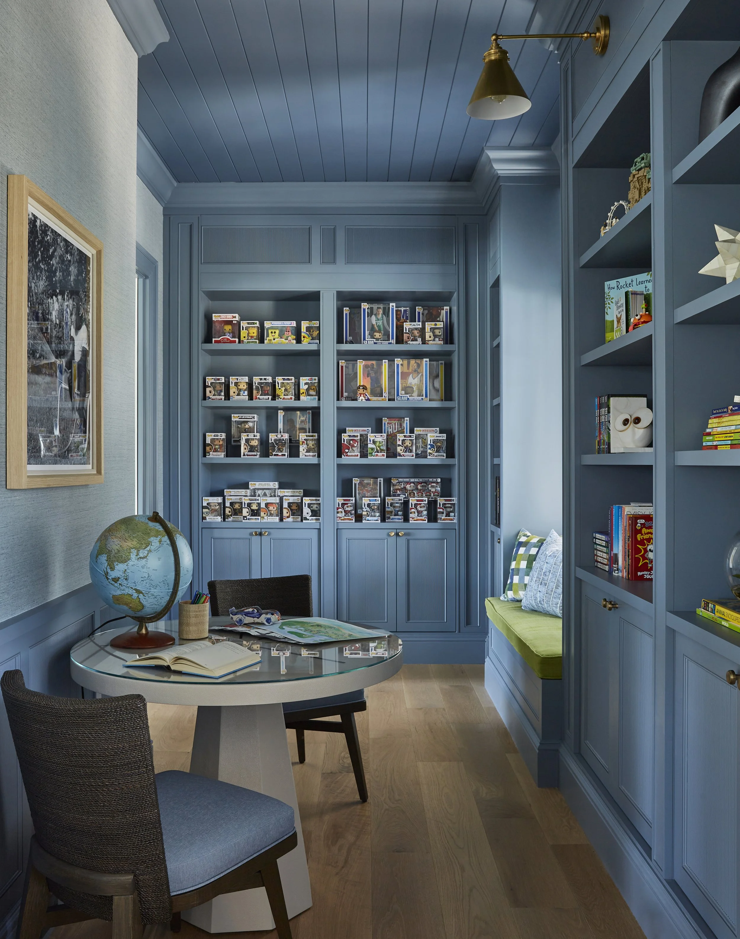A cozy reading nook with blue built-in shelves filled with books and collectibles, a window seat with green cushions, a round desk with a globe, open books, and a small container of pens, and two chairs, one upholstered and one wicker, in a room with