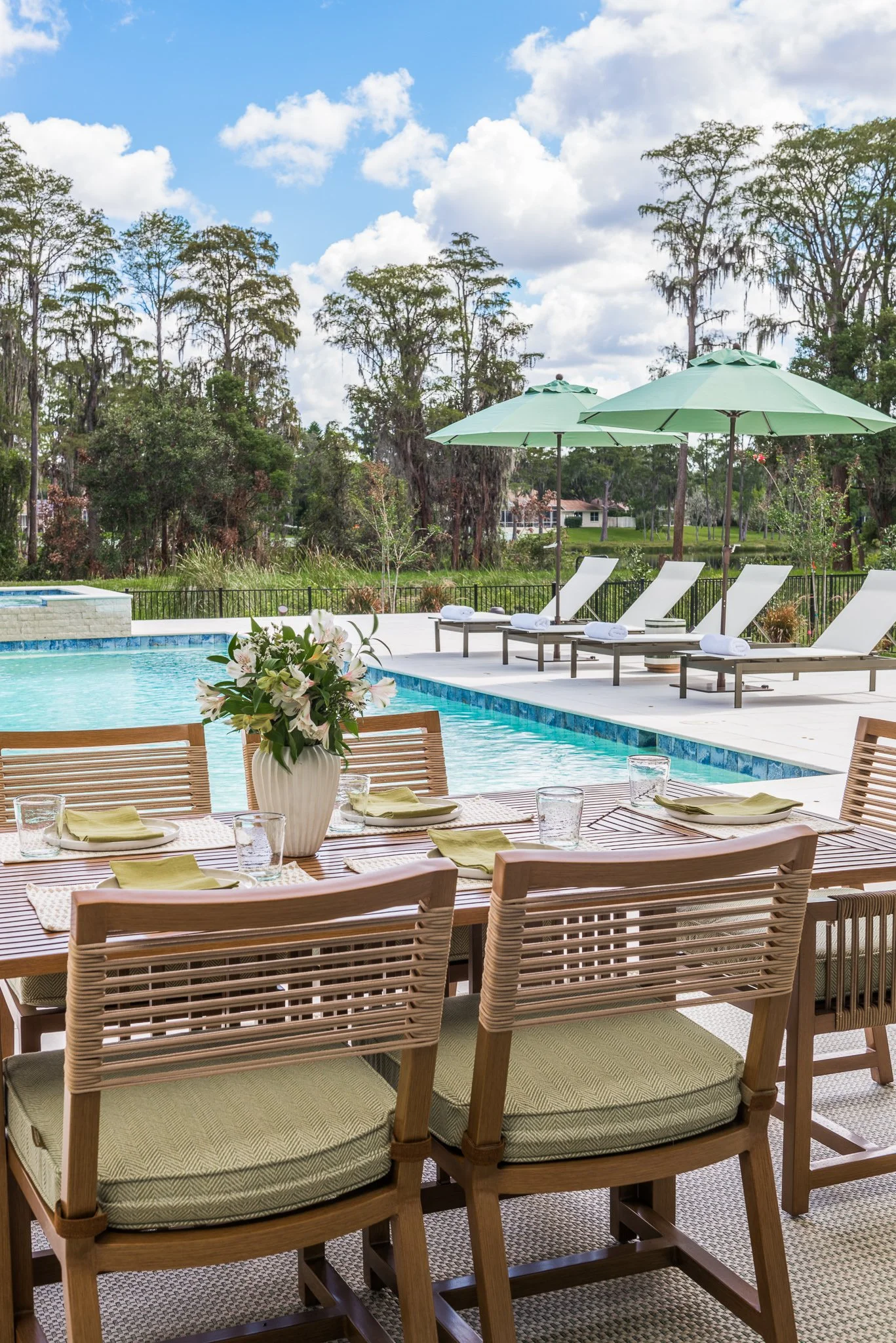 A poolside dining area with a wooden table set with plates, napkins, and glassware, next to a swimming pool with lounge chairs and large umbrellas, overlooking a green landscape with trees under a partly cloudy sky.