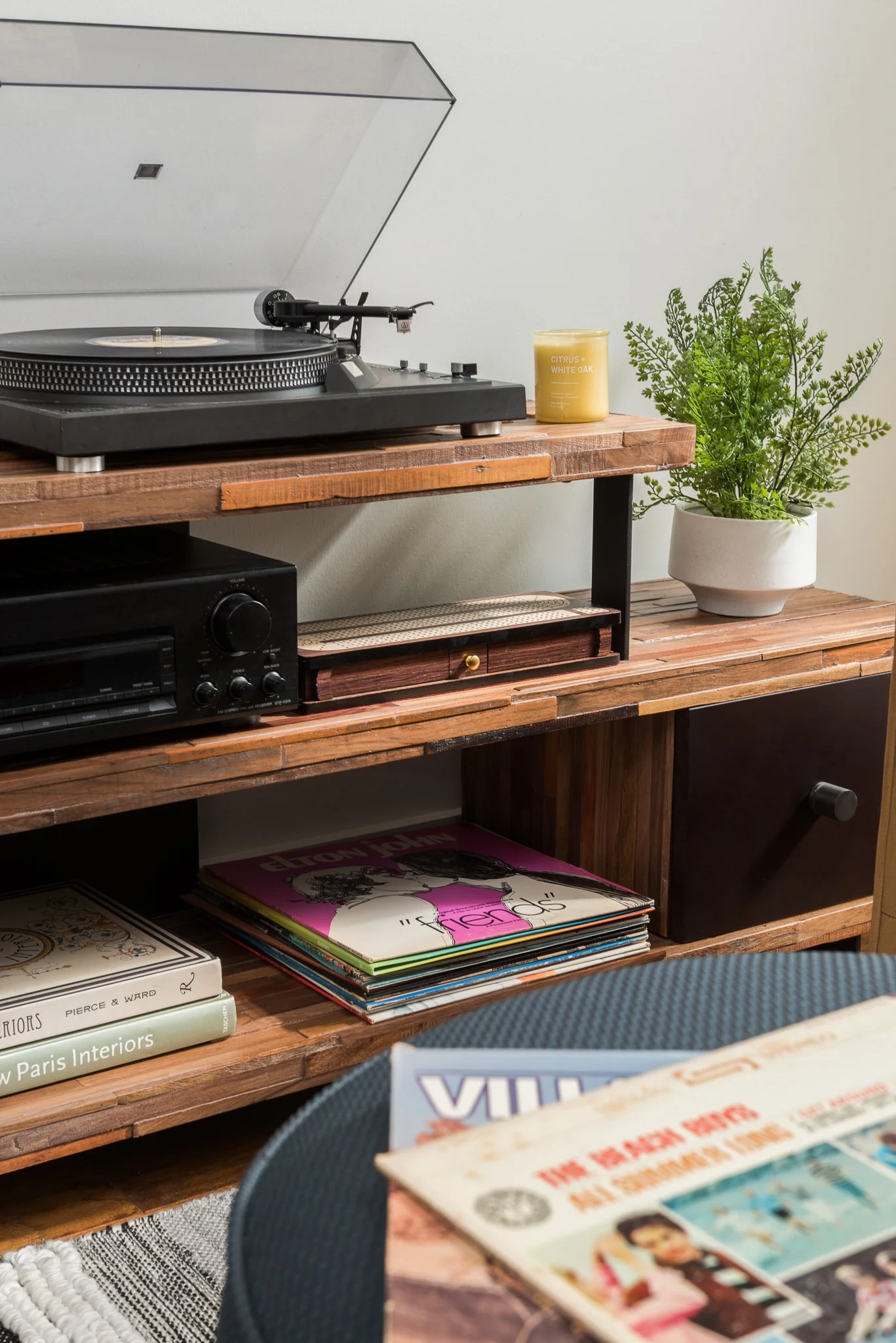 A wooden entertainment center with a turntable on top, a record player beneath, and shelves holding vinyl records, a black speaker, and decorative items including a candle and a potted plant.