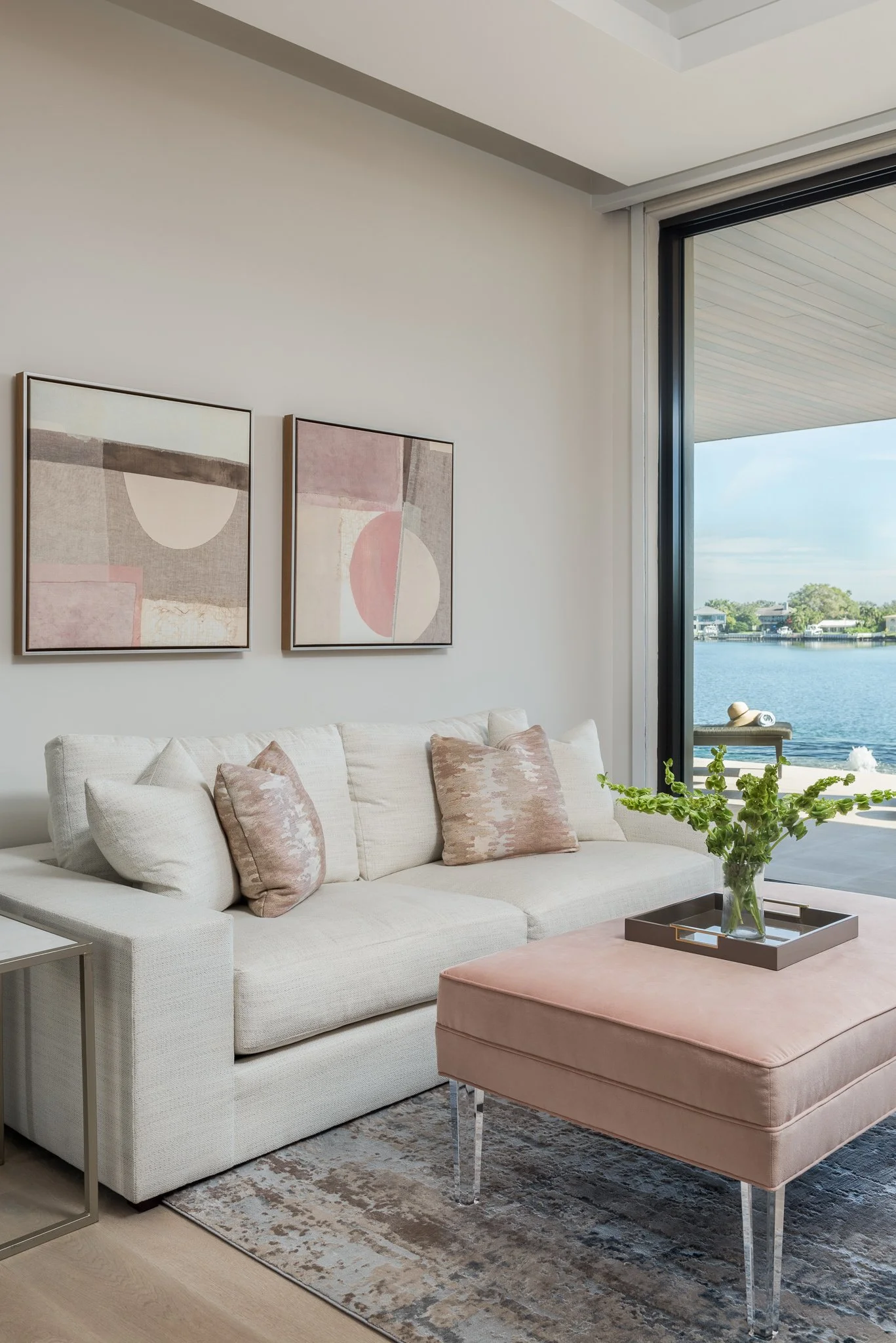 Modern living room with a white sofa, pink cushions, abstract artwork on the wall, a pink ottoman with a tray of green flowers, and large glass doors overlooking a lake.