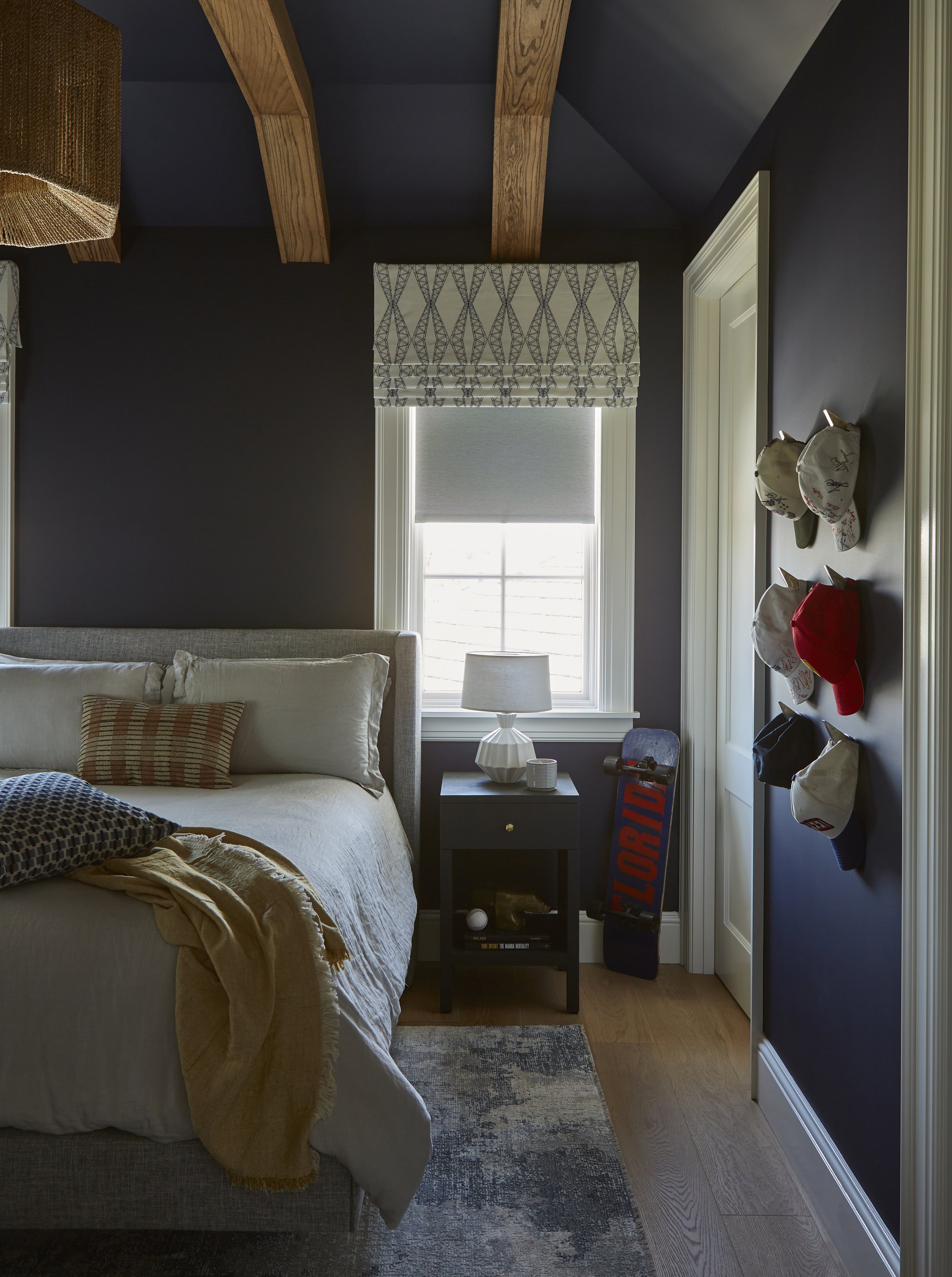 A bedroom with dark navy walls, wooden exposed ceiling beams, and a window with a patterned Roman shade. There is a bed with beige and patterned pillows, a nightstand with a white table lamp, and a skateboard with the word "FLORIDA" leaning against t