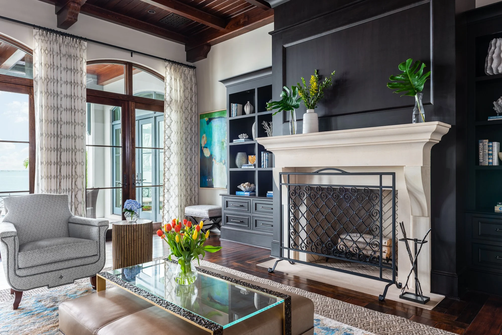 Living room with a fireplace, bookshelf, armchair, flowers, and large windows with curtains overlooking water.