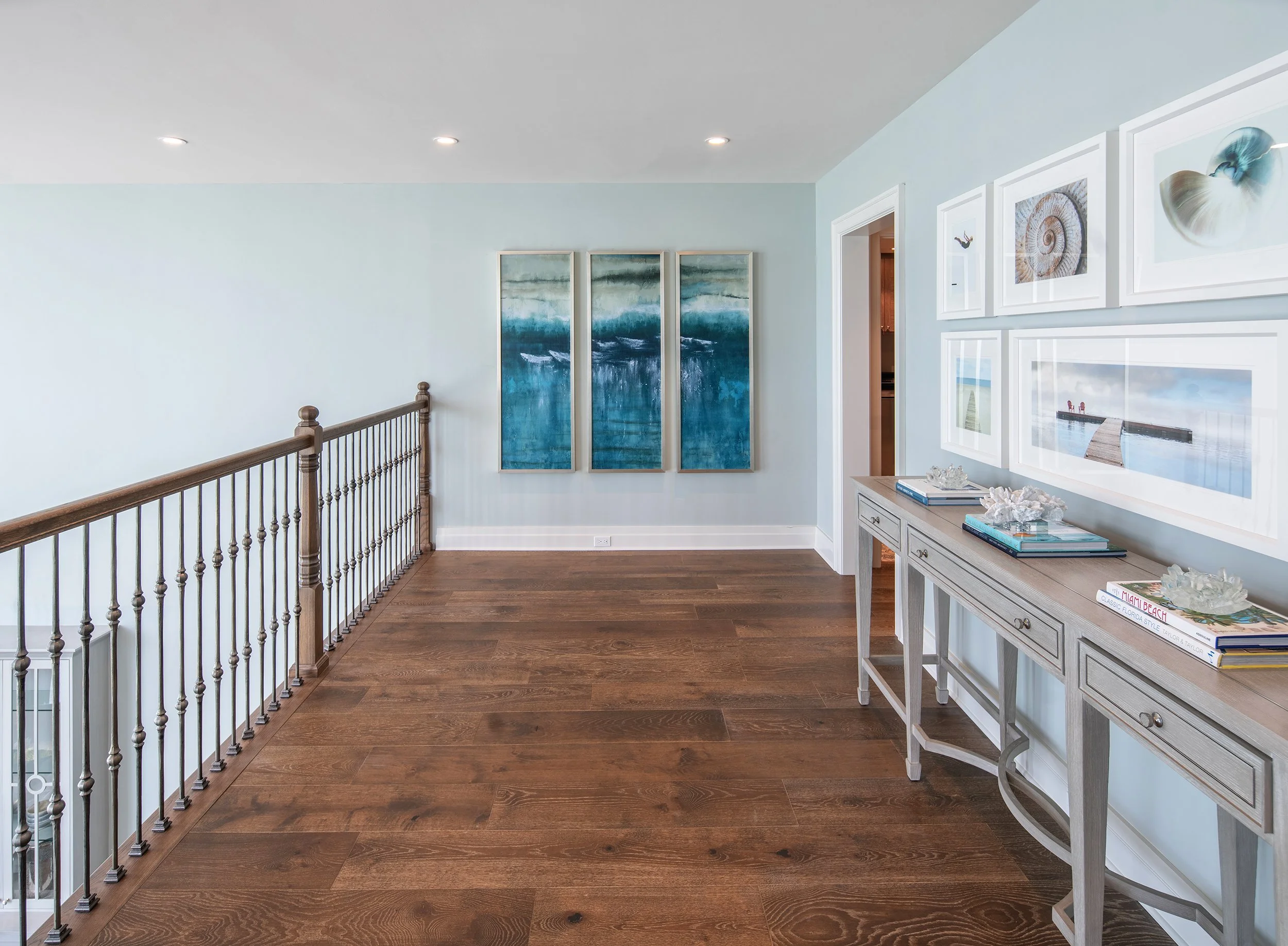 A hallway with wooden flooring, light blue walls, and white framed artwork, including coastal and beach themes, with a gray console table decorated with books and coral, and a staircase with wooden railing on the left.