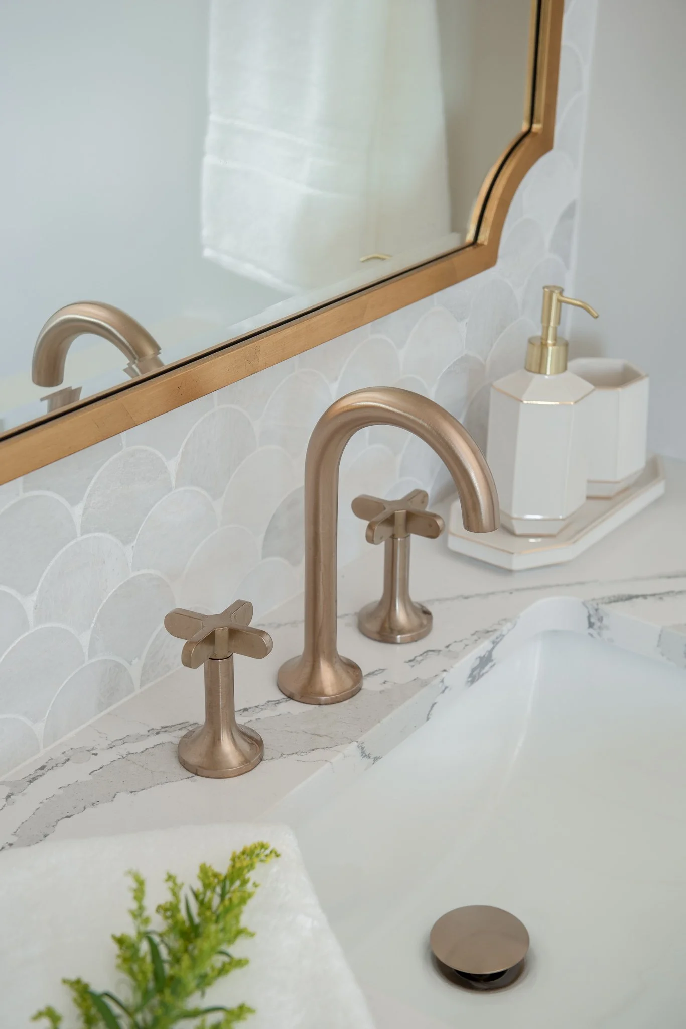Close-up of a bathroom countertop with a gold-toned faucet, matching cross-handle taps, a white hexagonal soap dispenser, and a large mirror reflecting white towels.