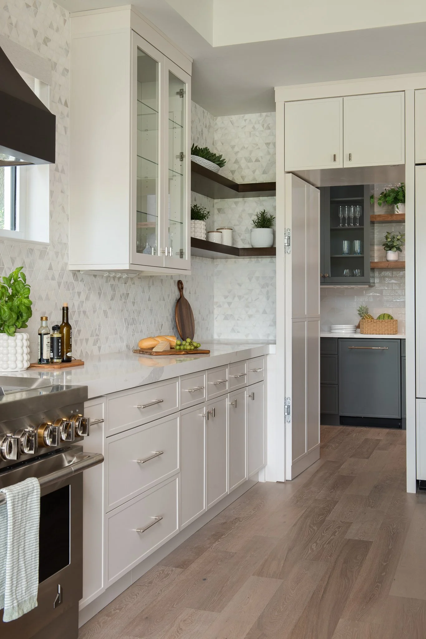 A modern kitchen with white cabinetry, wooden open shelves with plants, a patterned marble backsplash, a stainless steel stove, and light hardwood floors.