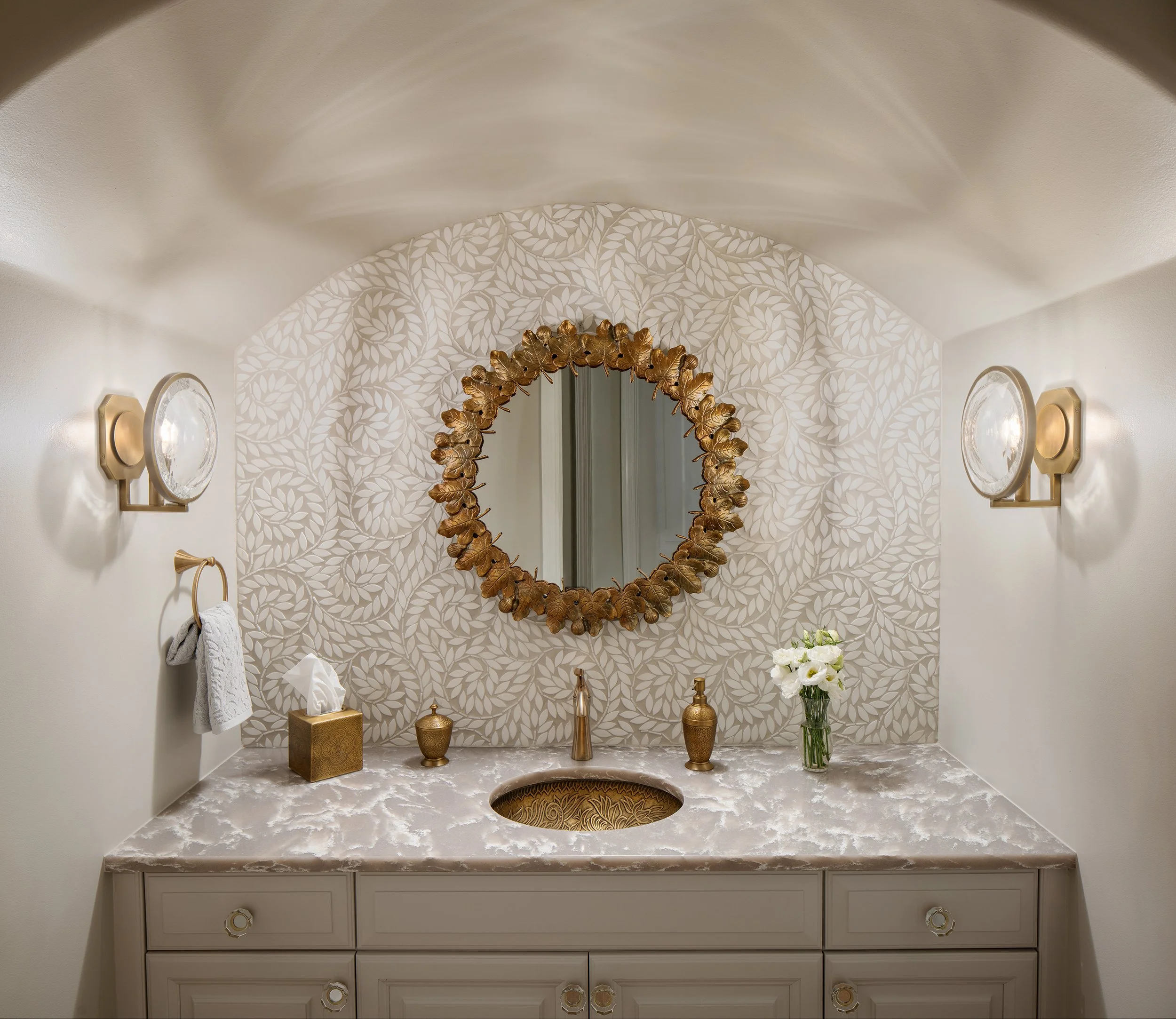 Elegant bathroom vanity with a gold-framed mirror, floral wallpaper, and decorative accessories including a tissue box, vases, and fresh flowers.
