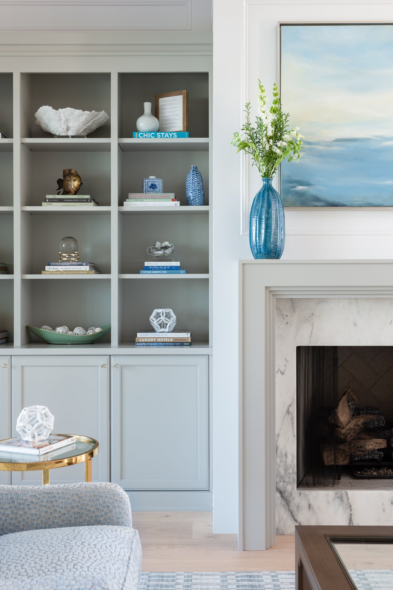 Living room with built-in shelves filled with decorative items, books, and vases. A blue glass vase with white flowers on a mantel with a marble fireplace, and a piece of abstract blue and white artwork.