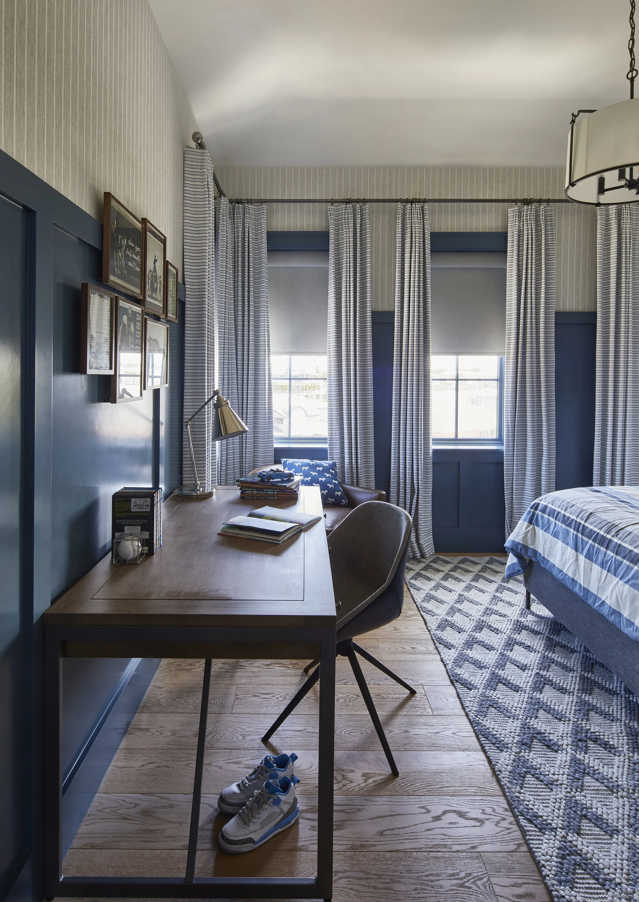 A cozy bedroom with a wooden desk, a chair, and a pair of sneakers underneath. There are two windows with striped curtains, a bed with blue and white bedding, and a patterned rug.