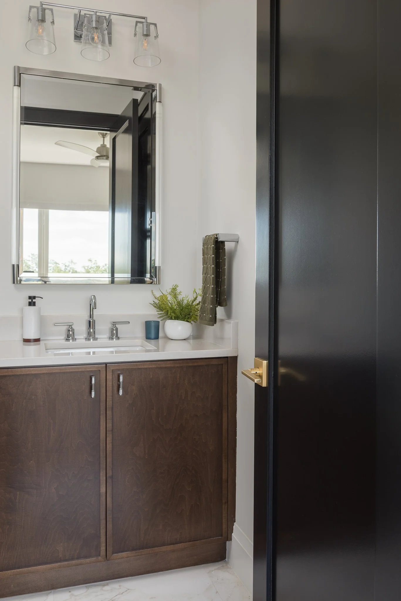 A bathroom vanity with a white countertop, a small sink, modern faucet, a mirror, a plant, a soap dispenser, and a small blue container. There is a window reflected in the mirror and a black door partially open to reveal the room.