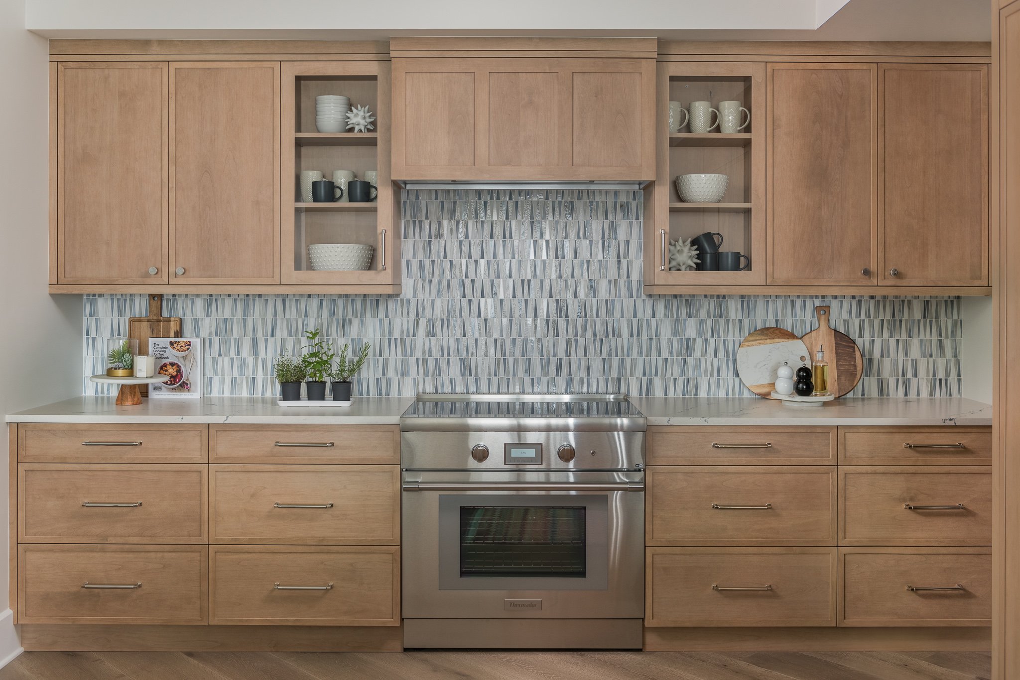 Kitchen with light wood cabinets, a stainless steel oven, and a geometric tile backsplash. Decor includes potted plants, plates, bowls, and kitchen accessories.