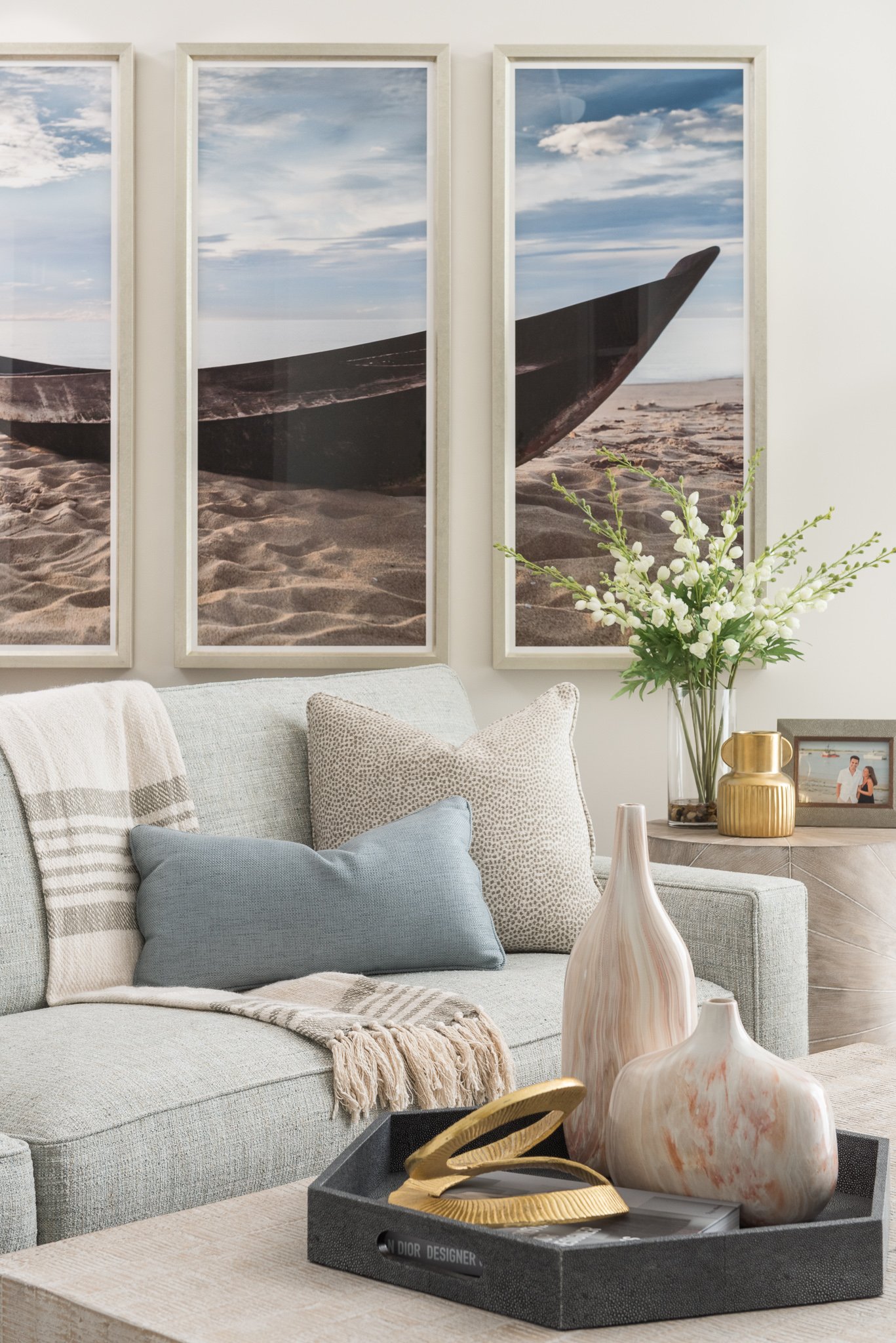 Living room with light gray sofa, throw pillows, and decorative vases, with three-panel wall art of a boat on a beach and clouds in the sky.