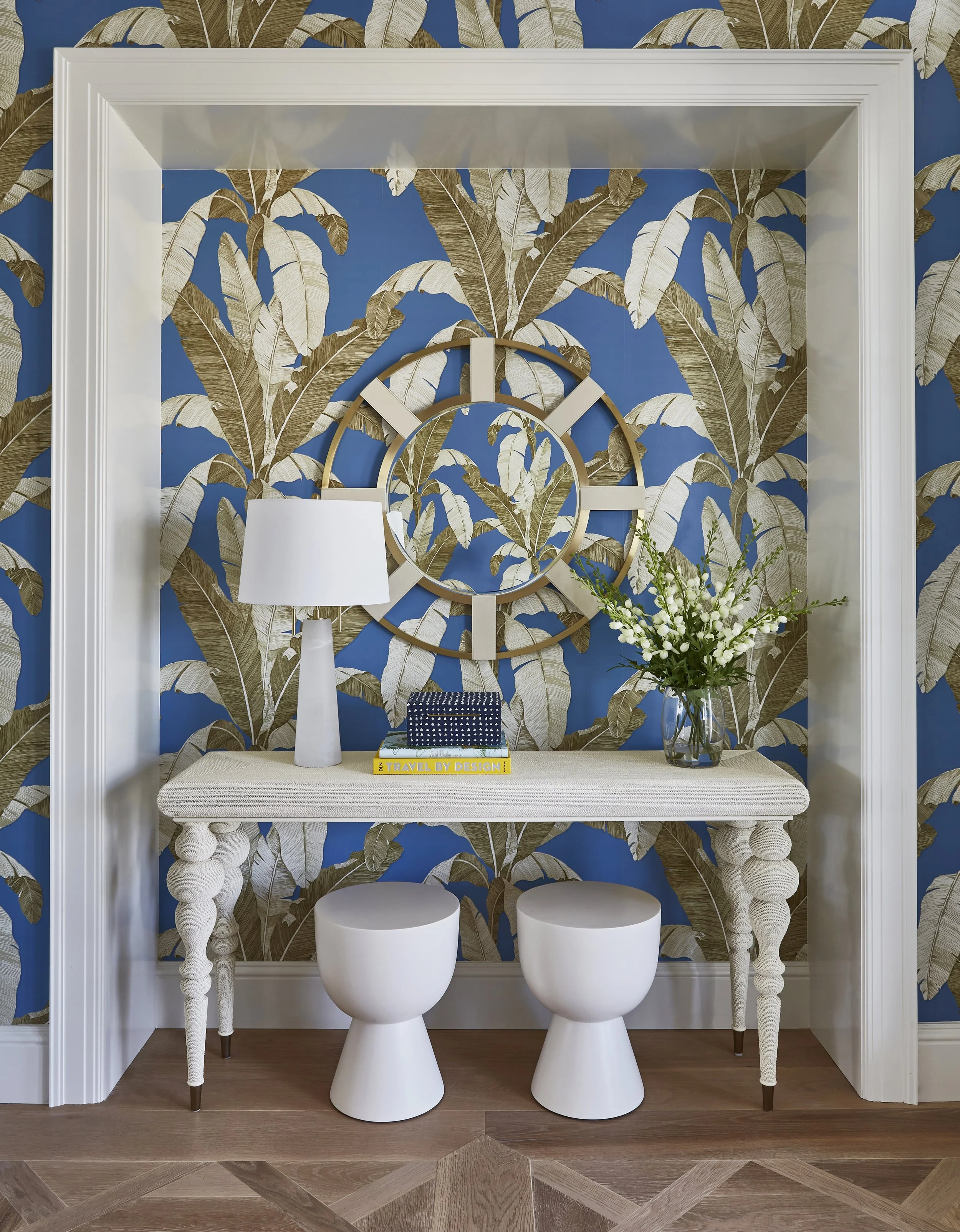 A decorative entryway nook with blue and beige botanical wallpaper, a large gold clock, a white table with textured legs, a white table lamp, a glass vase with white flowers, and two white stool-like seats.