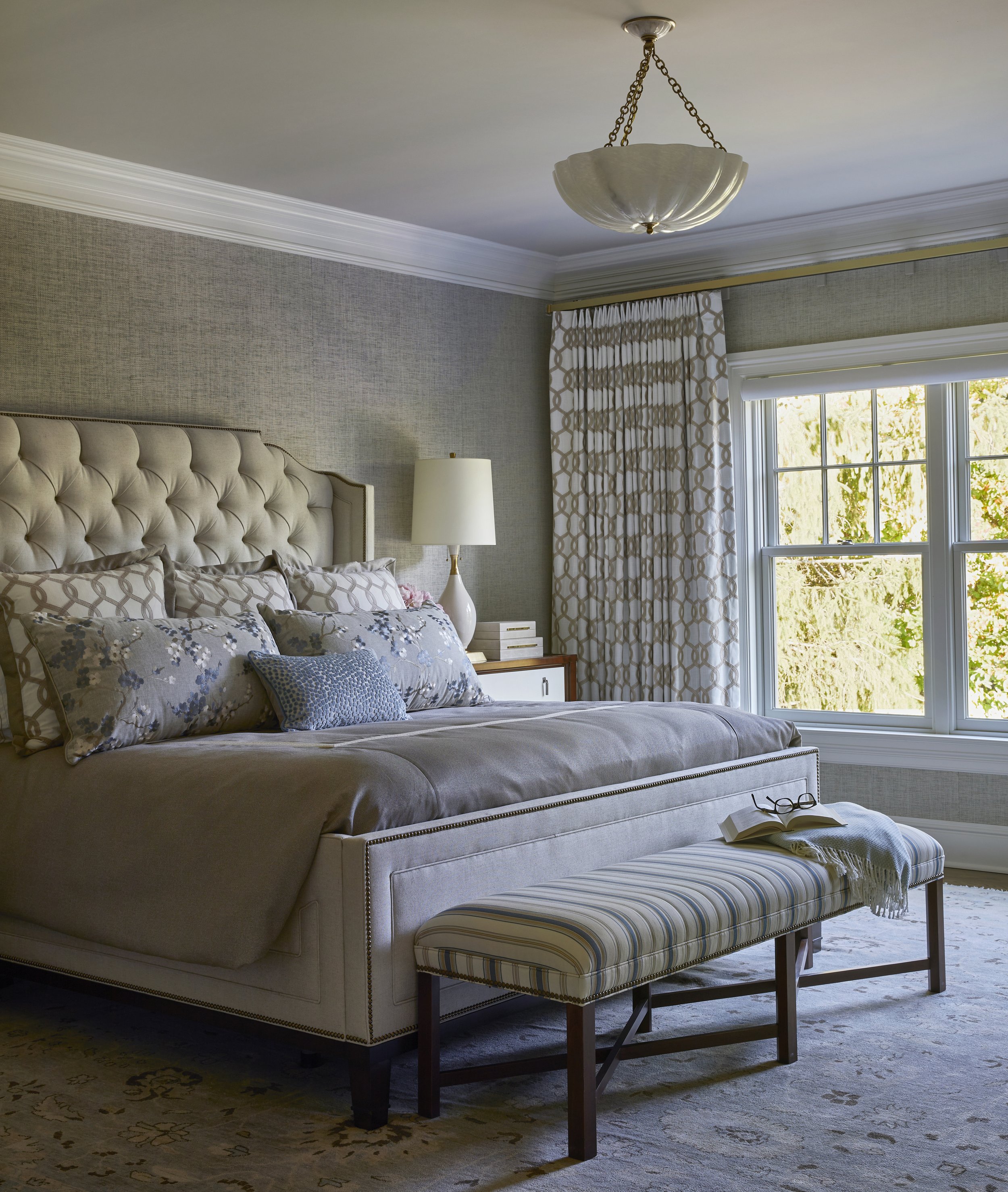 A bedroom with a tufted upholstered bed, decorative pillows, a bench with striped upholstery at the foot of the bed, window with patterned curtains, a nightstand with a white lamp, and a chandelier hanging from the ceiling.