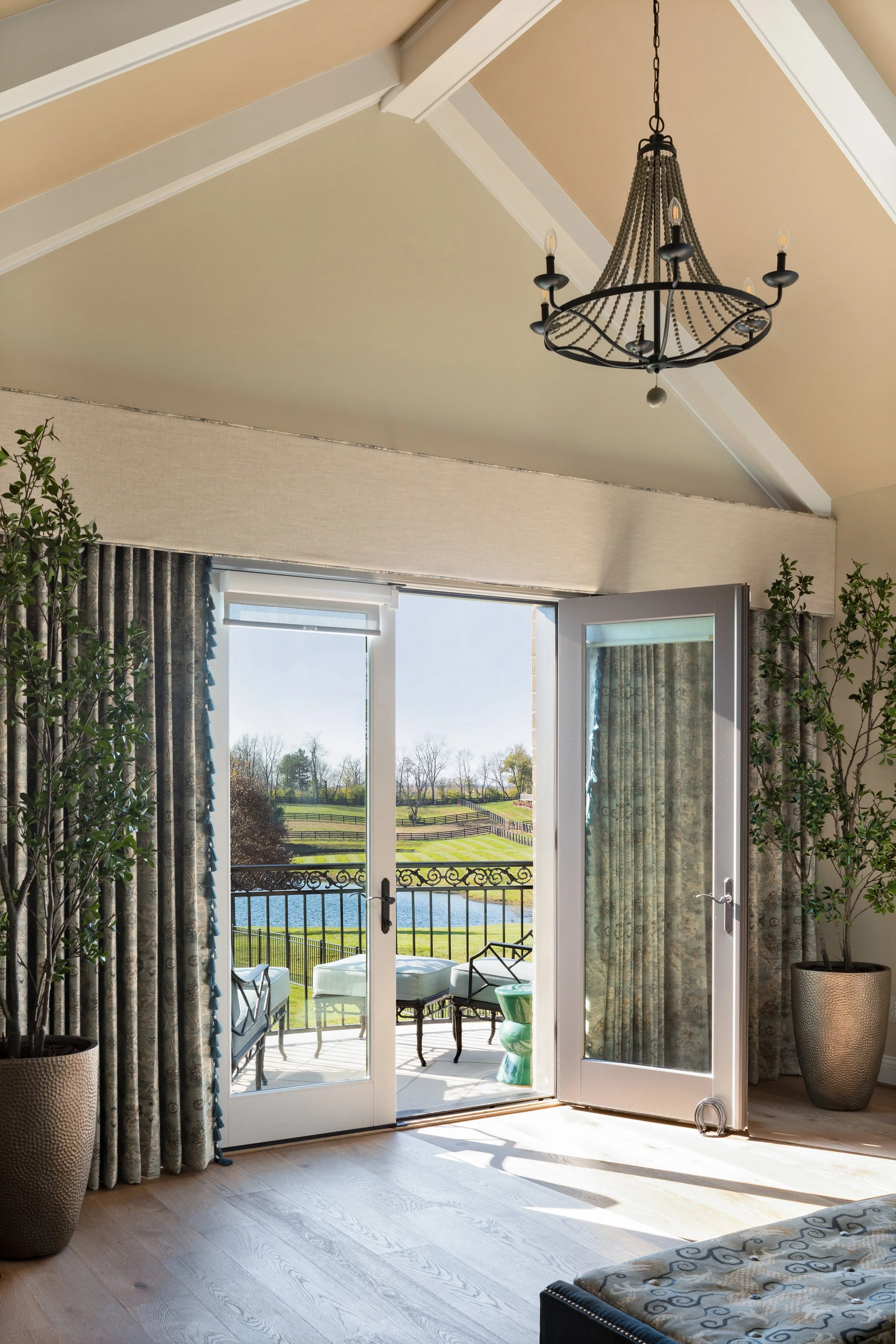 Sunlit living room with open glass doors leading to a balcony overlooking a pond and green landscape, decorated with potted plants, curtains, and a chandelier.