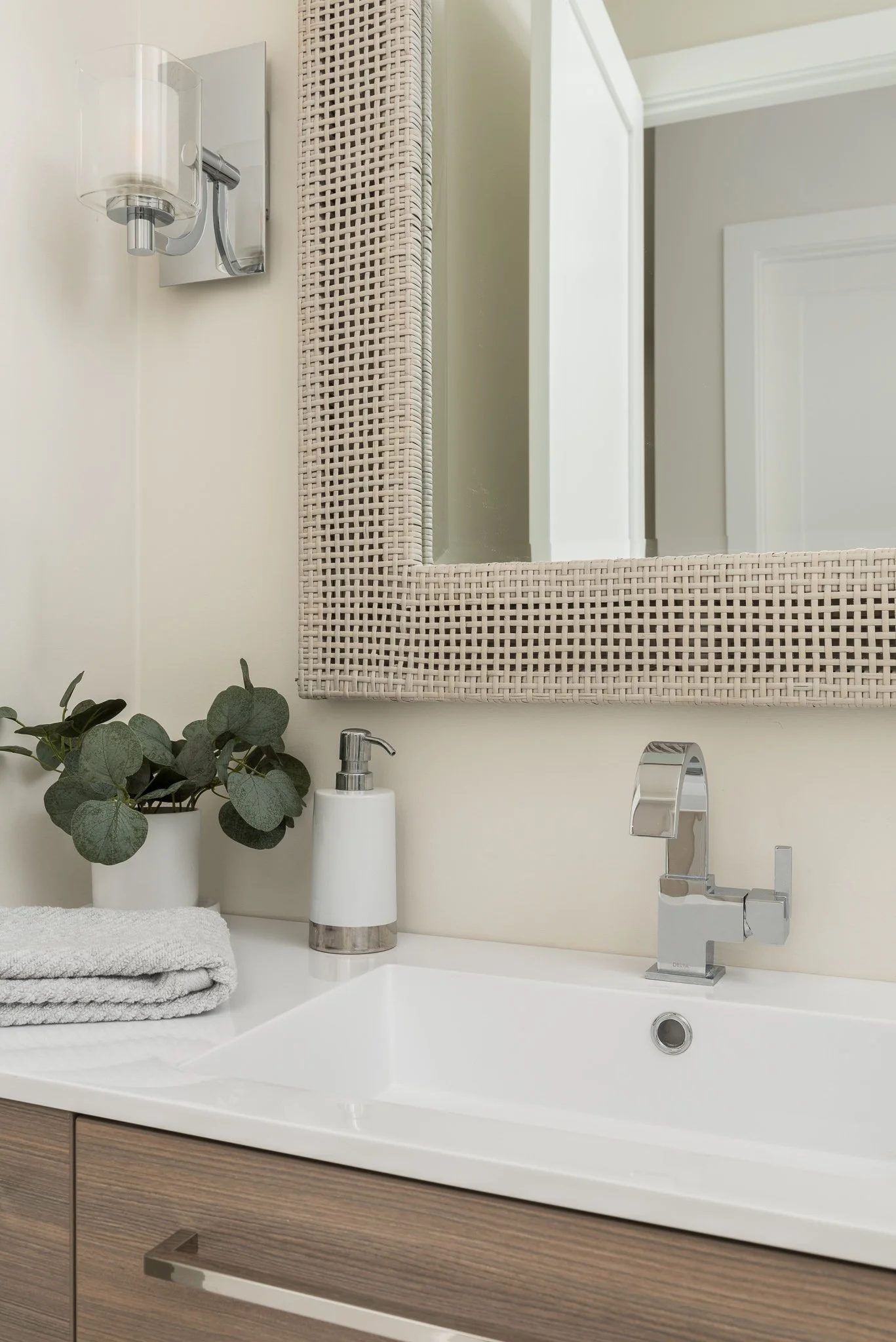 A bathroom sink with a white countertop, a modern chrome faucet, and a soap dispenser. A potted eucalyptus plant, a folded towel, and a mirror with a woven frame are also visible.