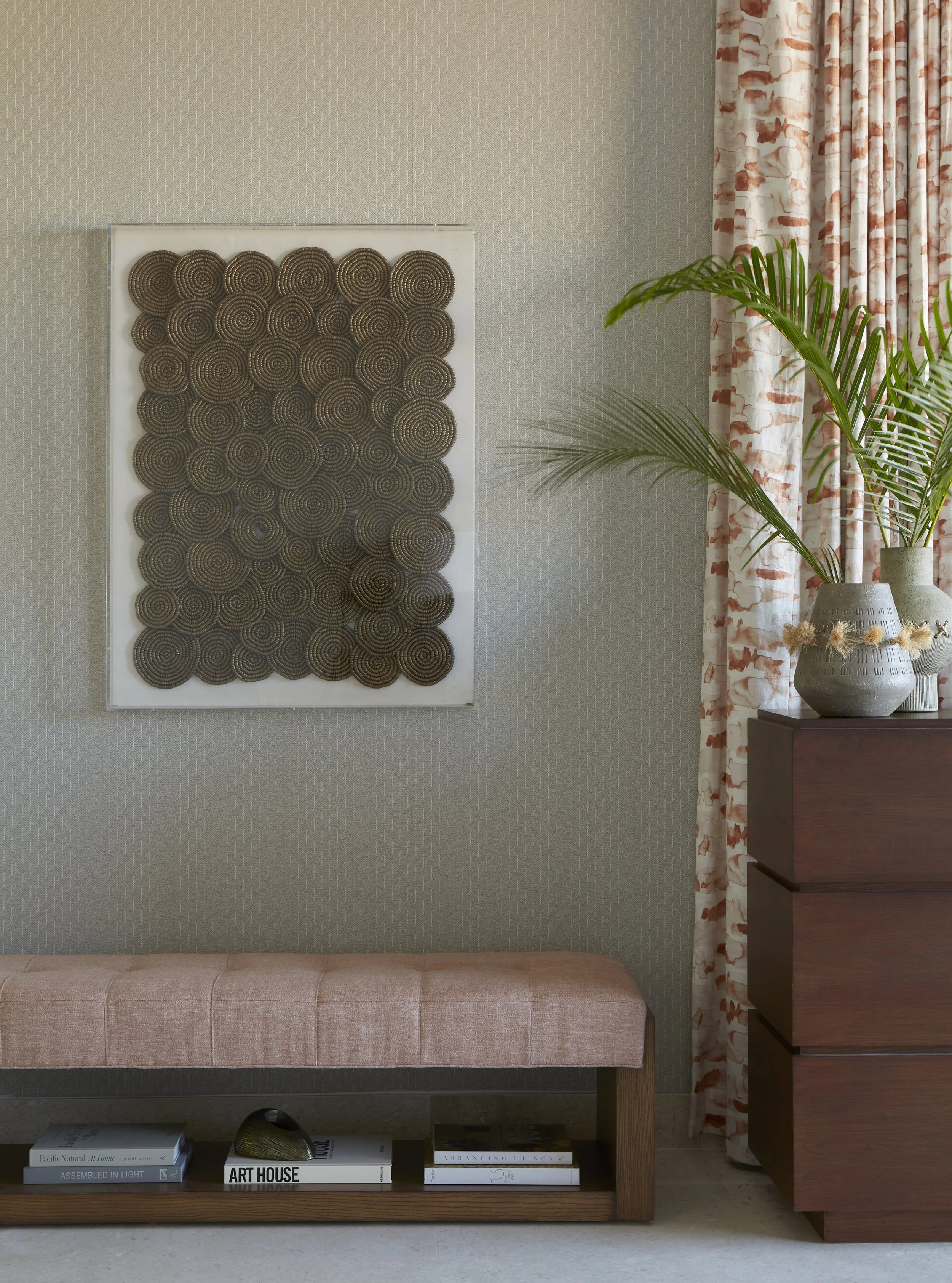 Interior of a room featuring textured gray wallpaper, a pink upholstered bench with a wooden base, a stack of books including one titled "Art House," and a wooden dresser topped with a large vase and tall green plants next to patterned curtains.