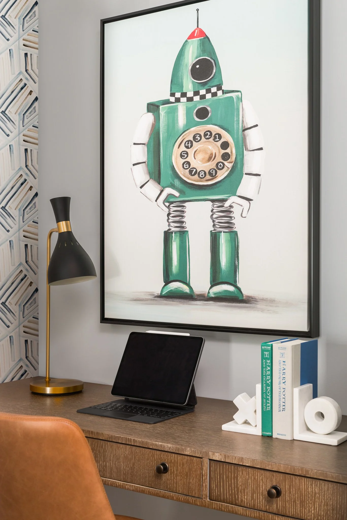 Large framed artwork of a retro robot with a rotary phone as its body, standing on a wooden desk next to a black and gold desk lamp, a tablet, and a small collection of Books and decorative objects.