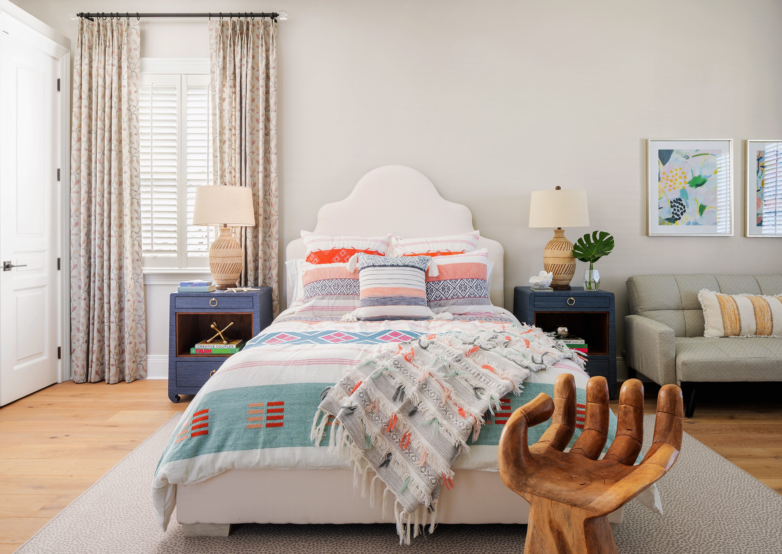 A bedroom with a white bed, colorful bedding, two blue nightstands with lamps, a beige sofa, framed wall art, and a unique wooden chair in the foreground.
