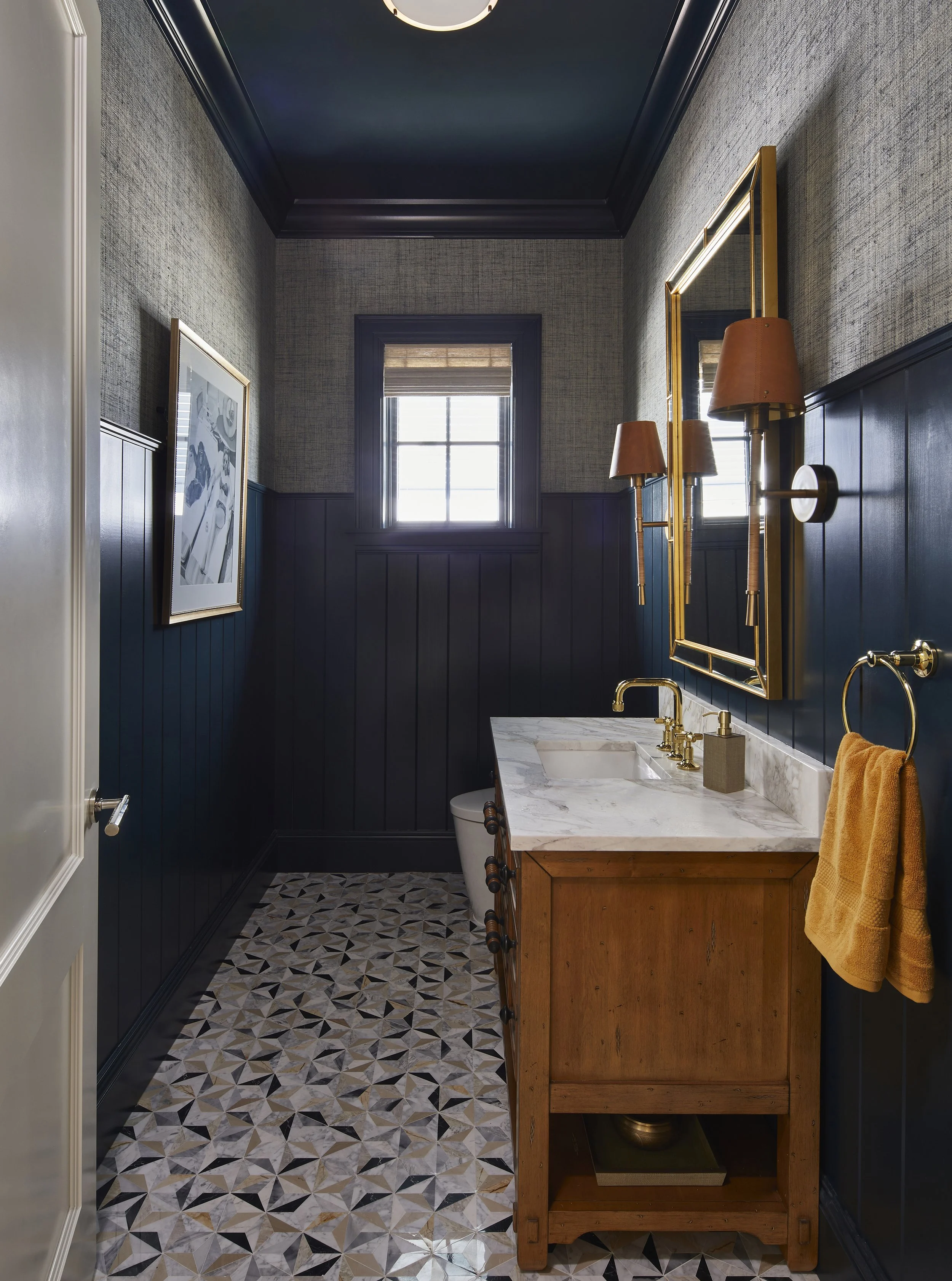 A small bathroom with dark blue wainscoting, textured wallpaper, a window with a shade, a wooden vanity with a marble top, gold fixtures, a yellow towel, and a geometric patterned tile floor.