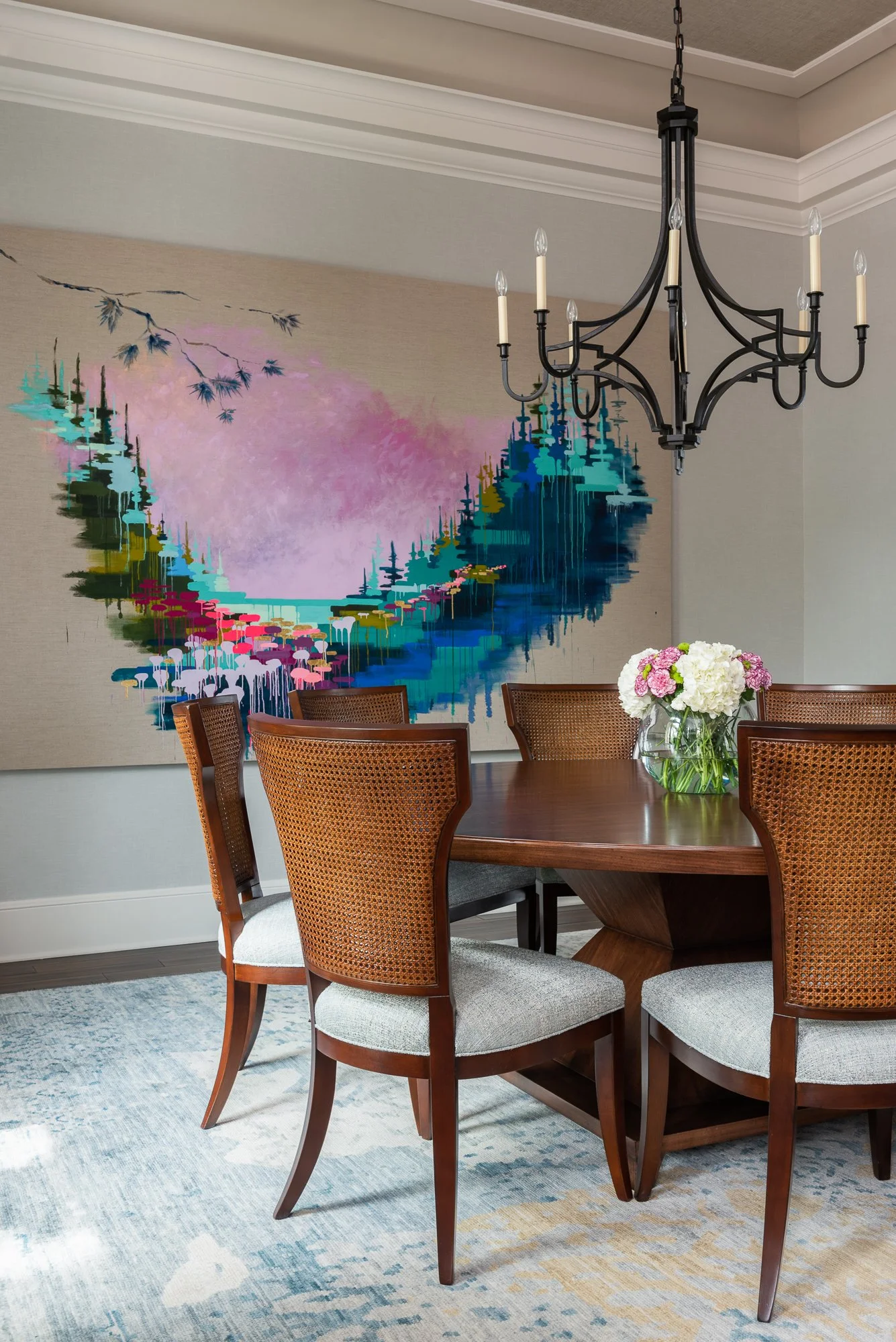 A dining room with a round wooden table, six chairs with woven backs, a floral centerpiece with white and pink flowers, a modern chandelier with multiple candle lights, a large abstract wall art depicting a colorful landscape, and a light-colored rug