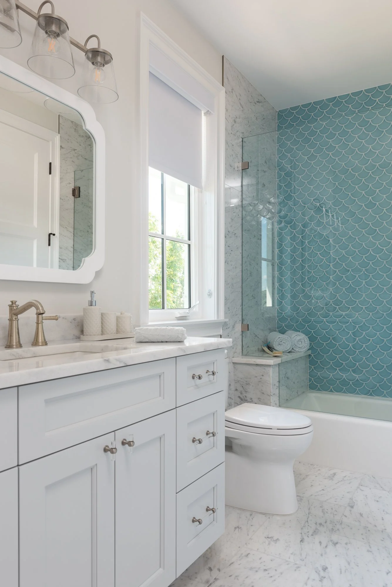 A modern bathroom with white cabinetry, a marble countertop, a mirror, a glass light fixture, a window with a white blind, a toilet, and a bathtub with a glass shower enclosure and blue fish scale tiles.