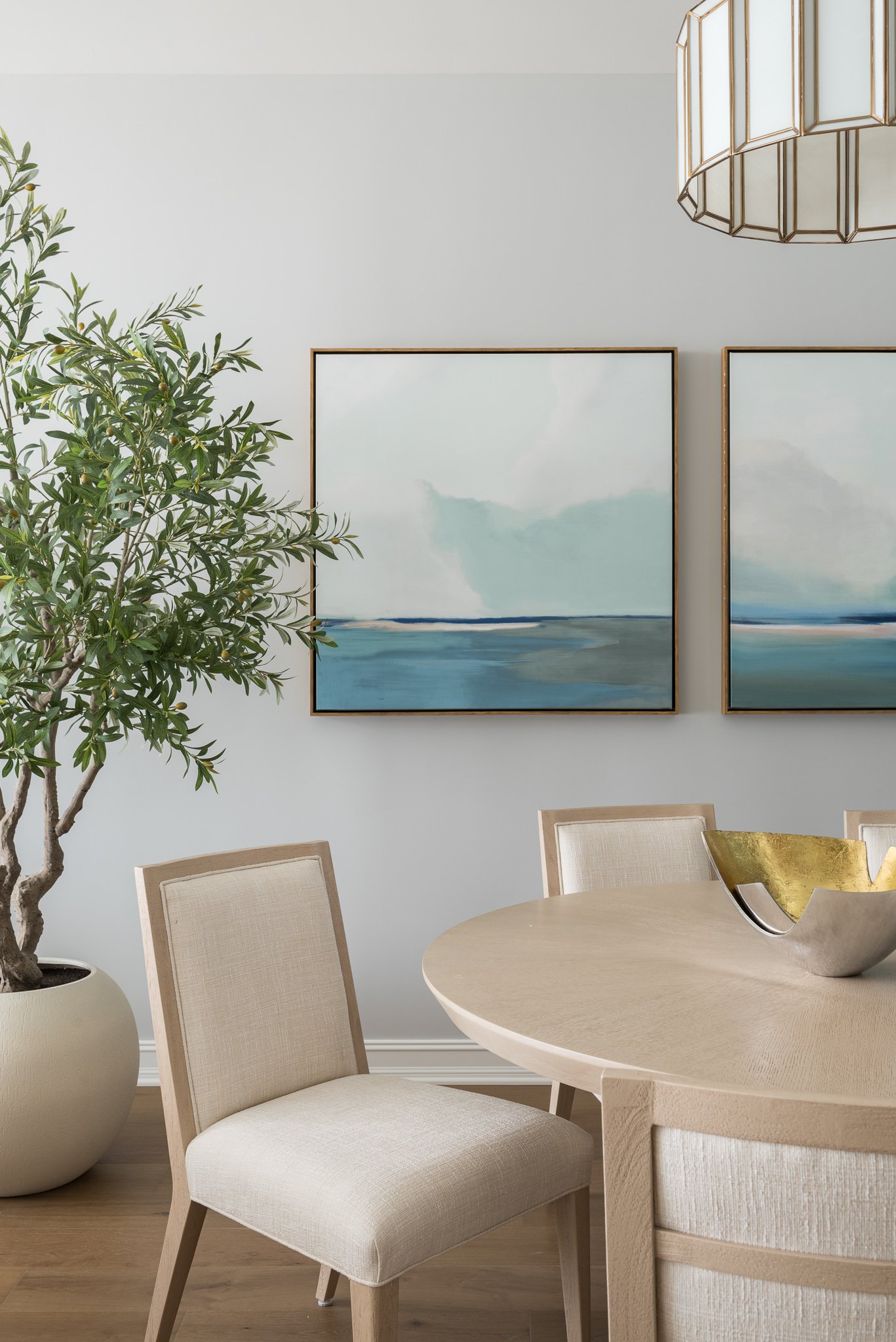 A modern dining room with a light wood round table, beige upholstered chairs, a large potted green plant, abstract seaside artwork on a white wall, and a geometric chandelier.