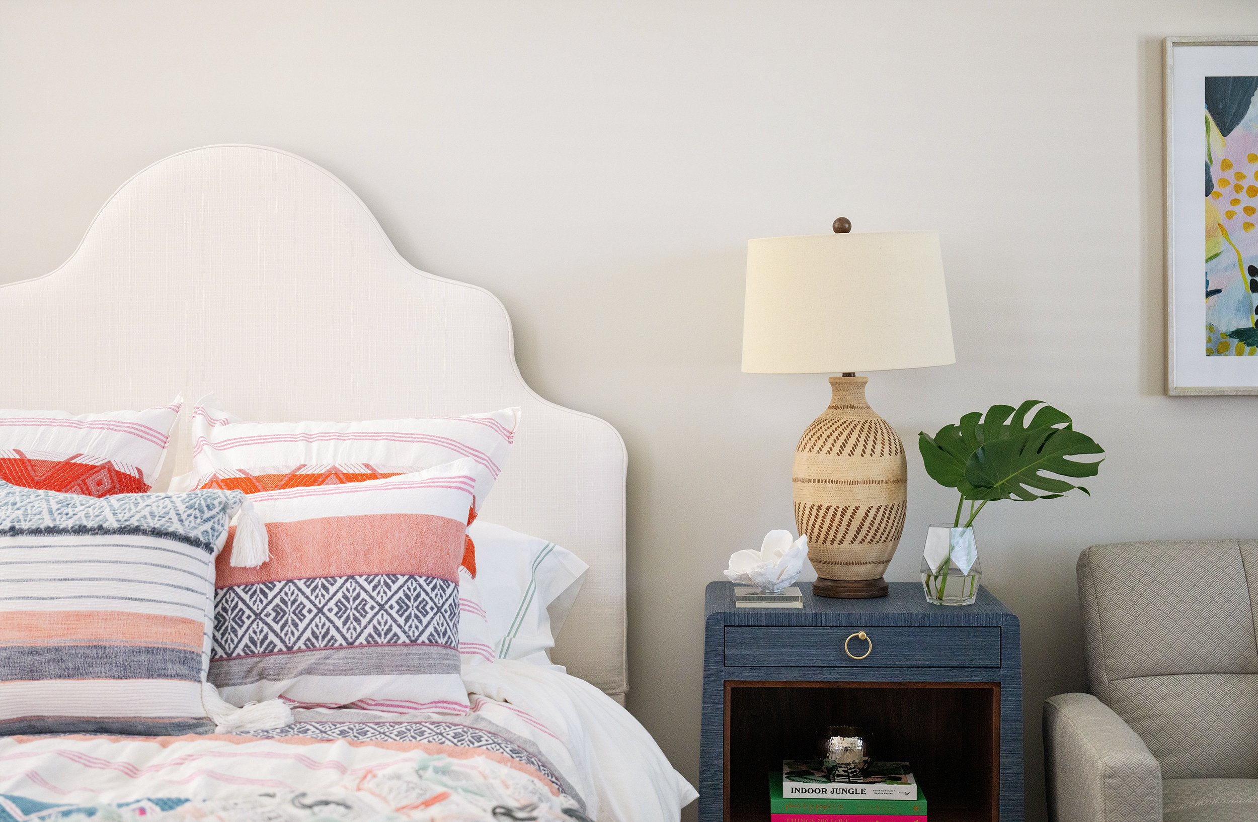 A bedroom with a white upholstered headboard, colorful pillows, a blue nightstand with a ceramic lamp, a large green leaf plant in a glass vase, and a framed colorful artwork on the wall.