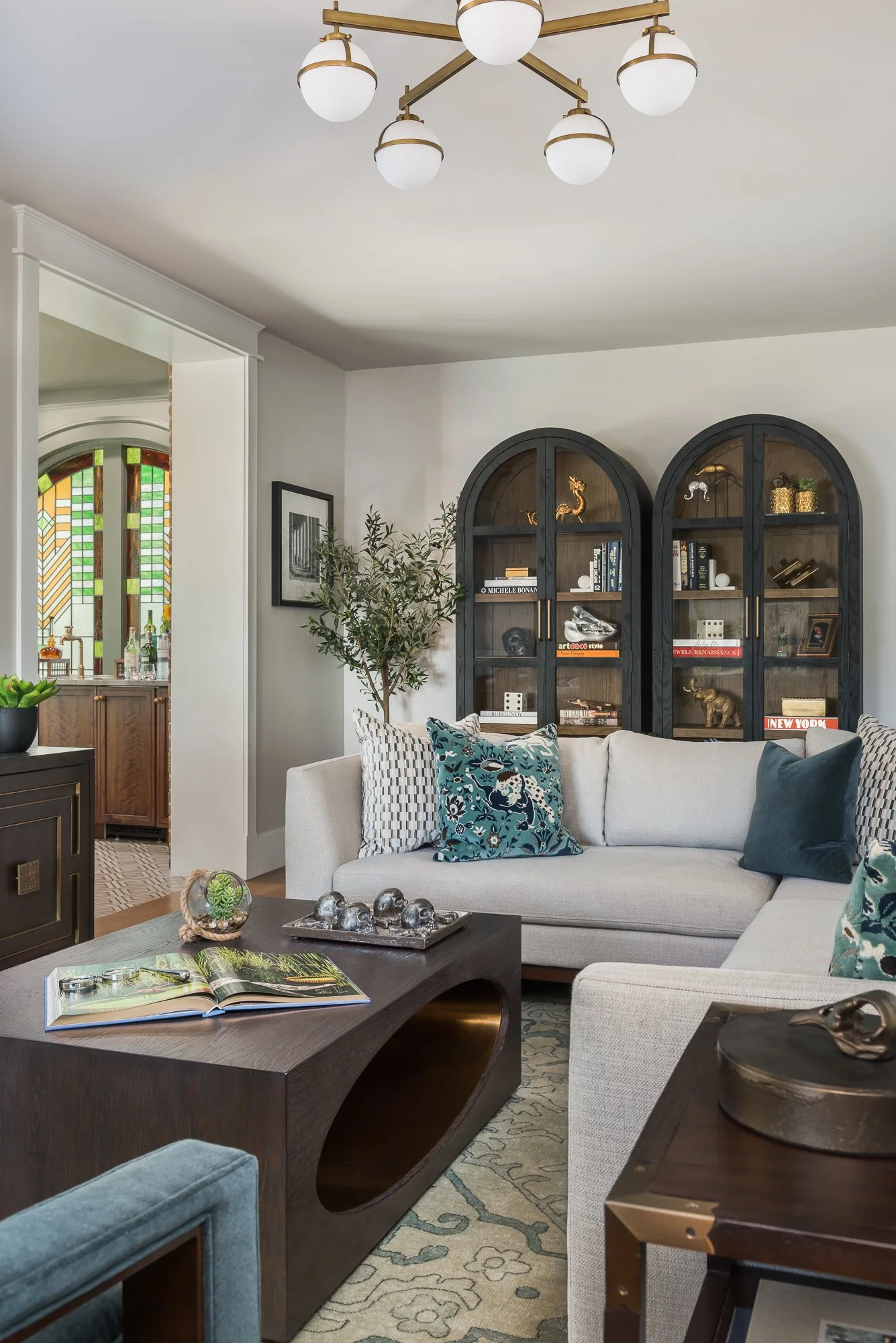 Living room with white sectional sofa, patterned and solid throw pillows, a wooden side table, dark wood bookshelf with decorative items, and a modern chandelier.