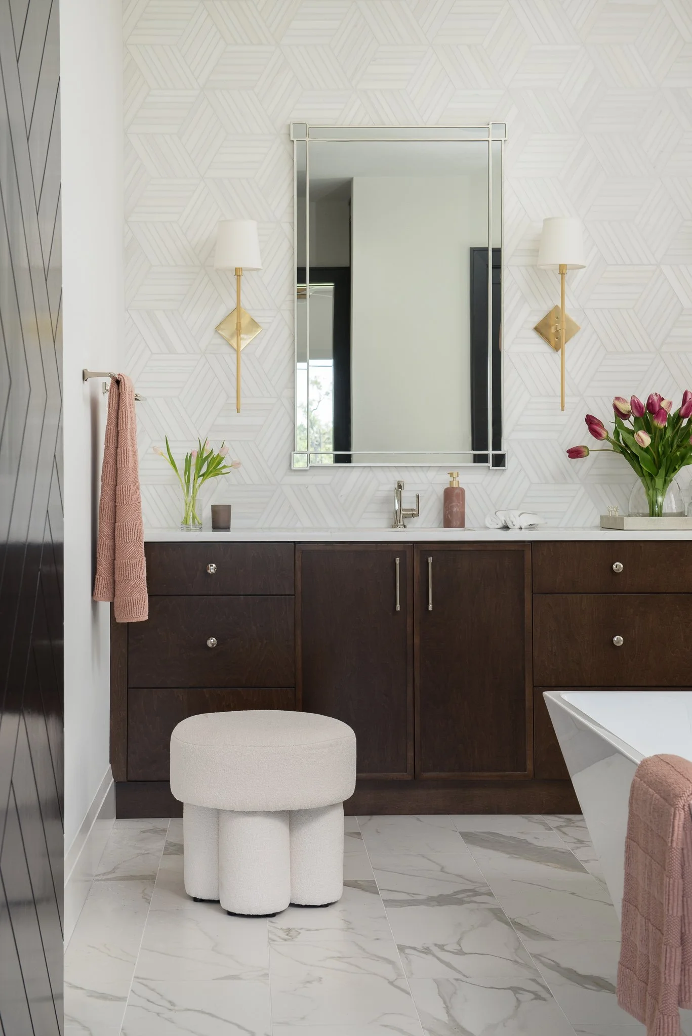A modern bathroom with a large mirror above a dark wood vanity, decorated with pink tulips and a vase, and flanked by two wall-mounted lamps, with marble tile flooring.