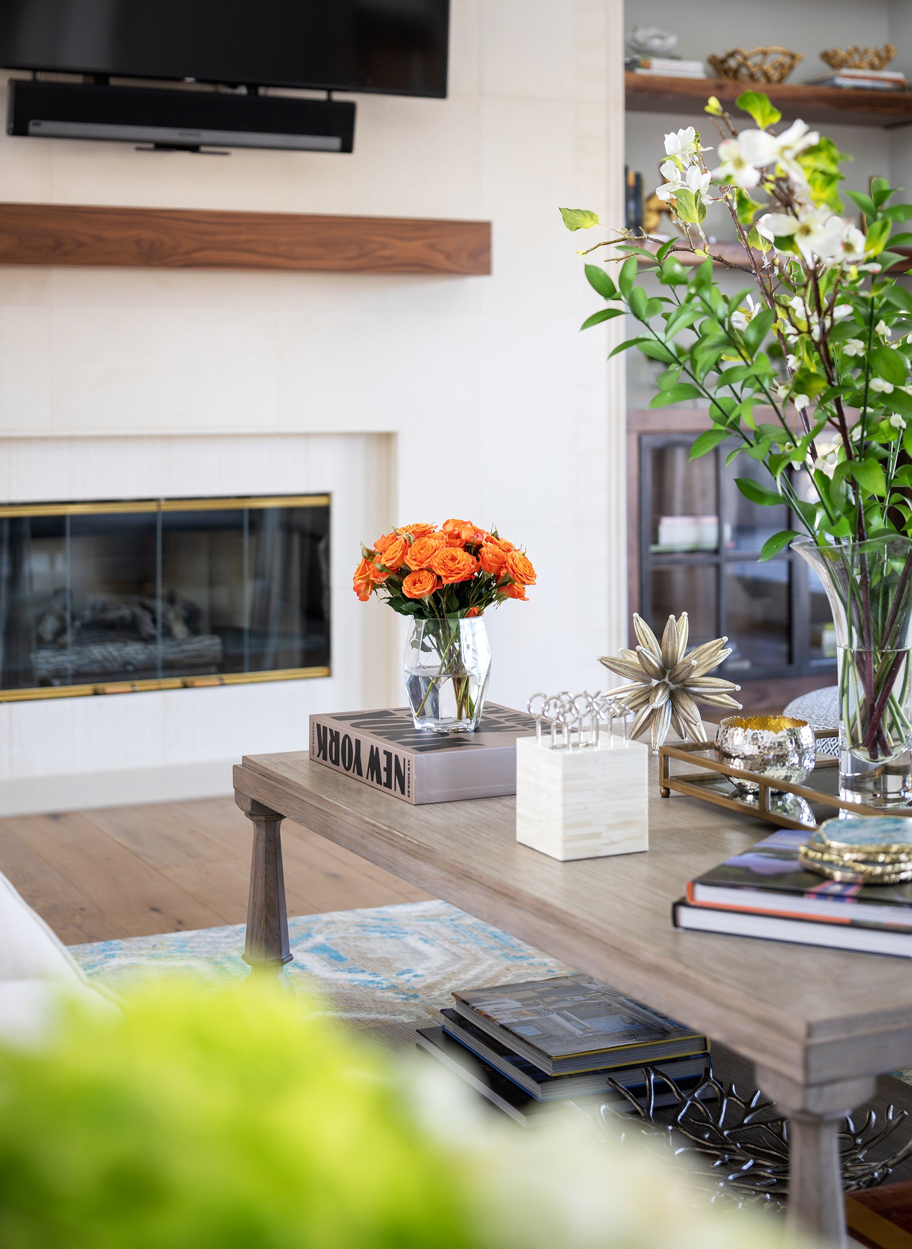 A cozy living room with a wooden table decorated with a bouquet of orange flowers in a glass vase, a stack of books, and decorative objects, with a fireplace, a TV mounted above, and shelves with various items in the background.