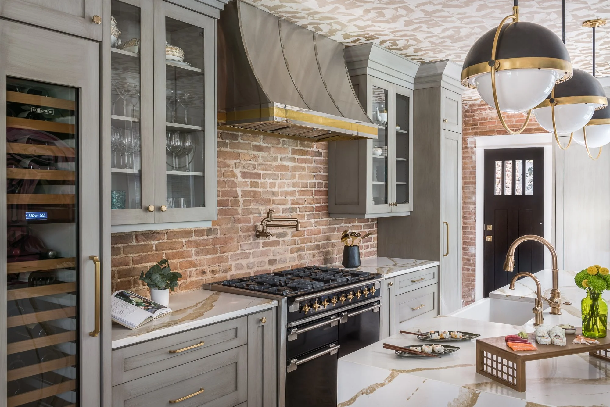 Modern kitchen with gray cabinetry, brick wall, gold accents, white marble countertops, and a black door.