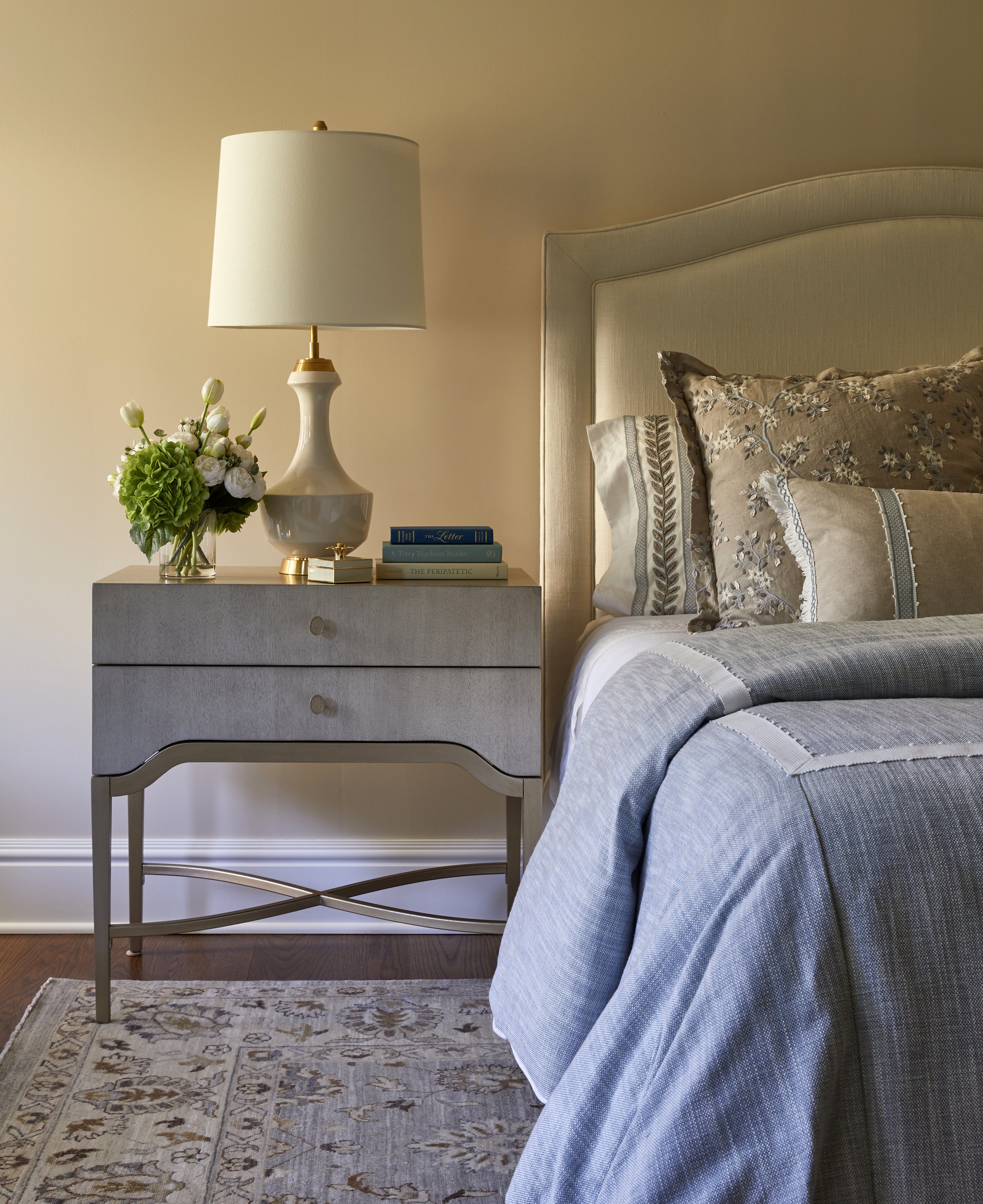 A bedroom with a gray nightstand, a white lamp, a glass vase with green flowers, a few books, a beige tufted headboard, and decorative pillows on the bed.