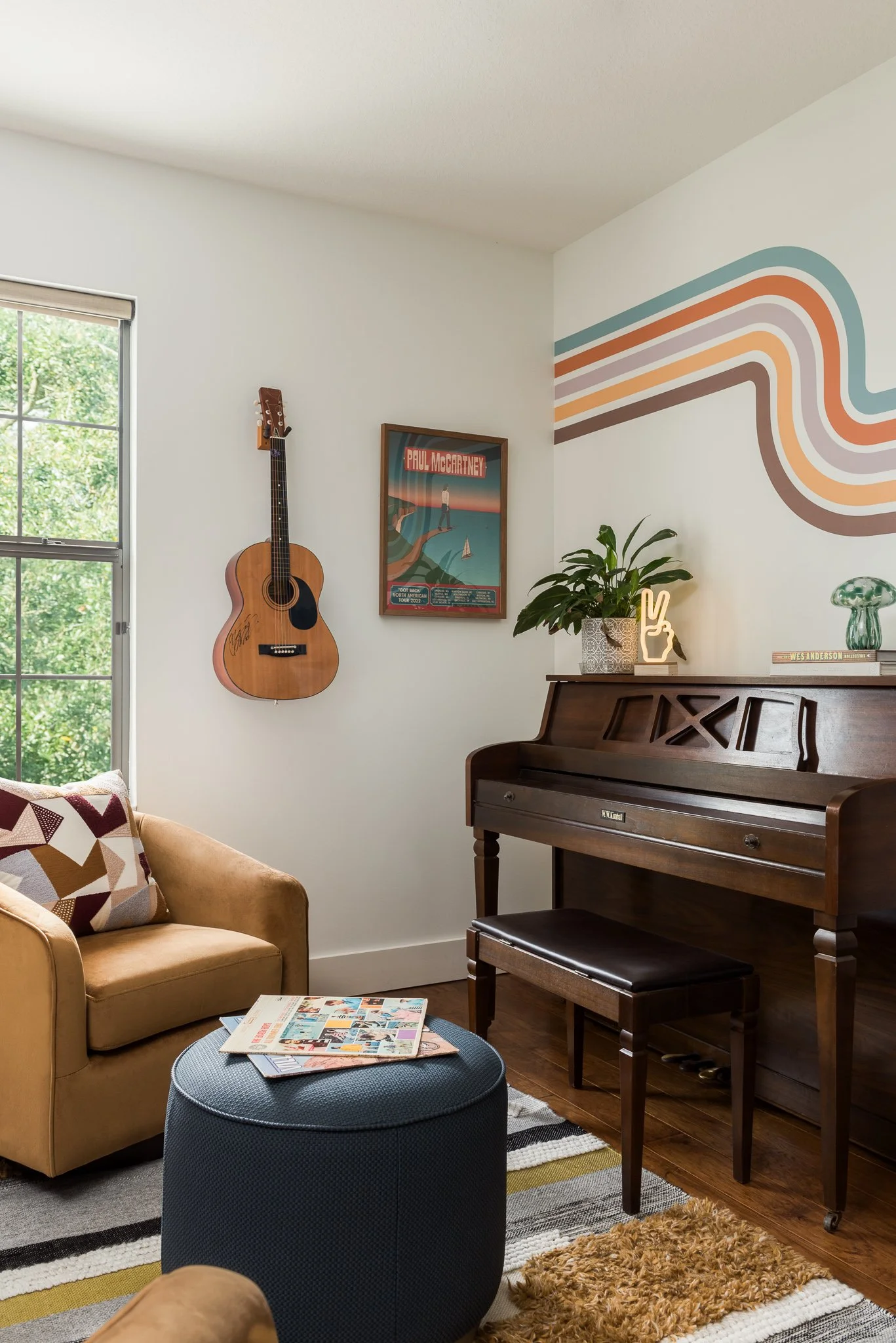A cozy living room corner with a beige sofa and patterned pillow, a dark blue ottoman with magazines, a wooden piano with decorative items and a plant on top, a framed Paul McCartney poster, a wall-mounted acoustic guitar, a window with green foliage