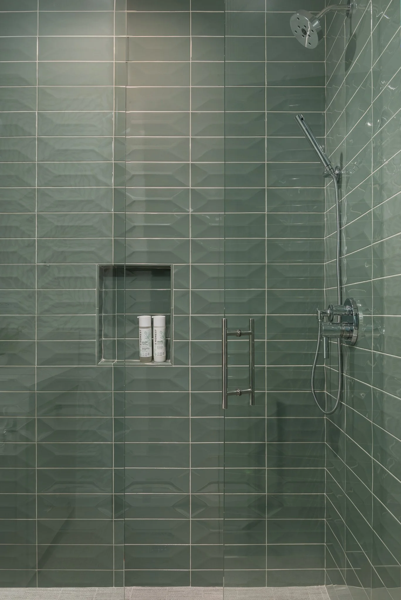 A modern walk-in shower with green tiled walls, a built-in niche with toiletries, and a glass door.