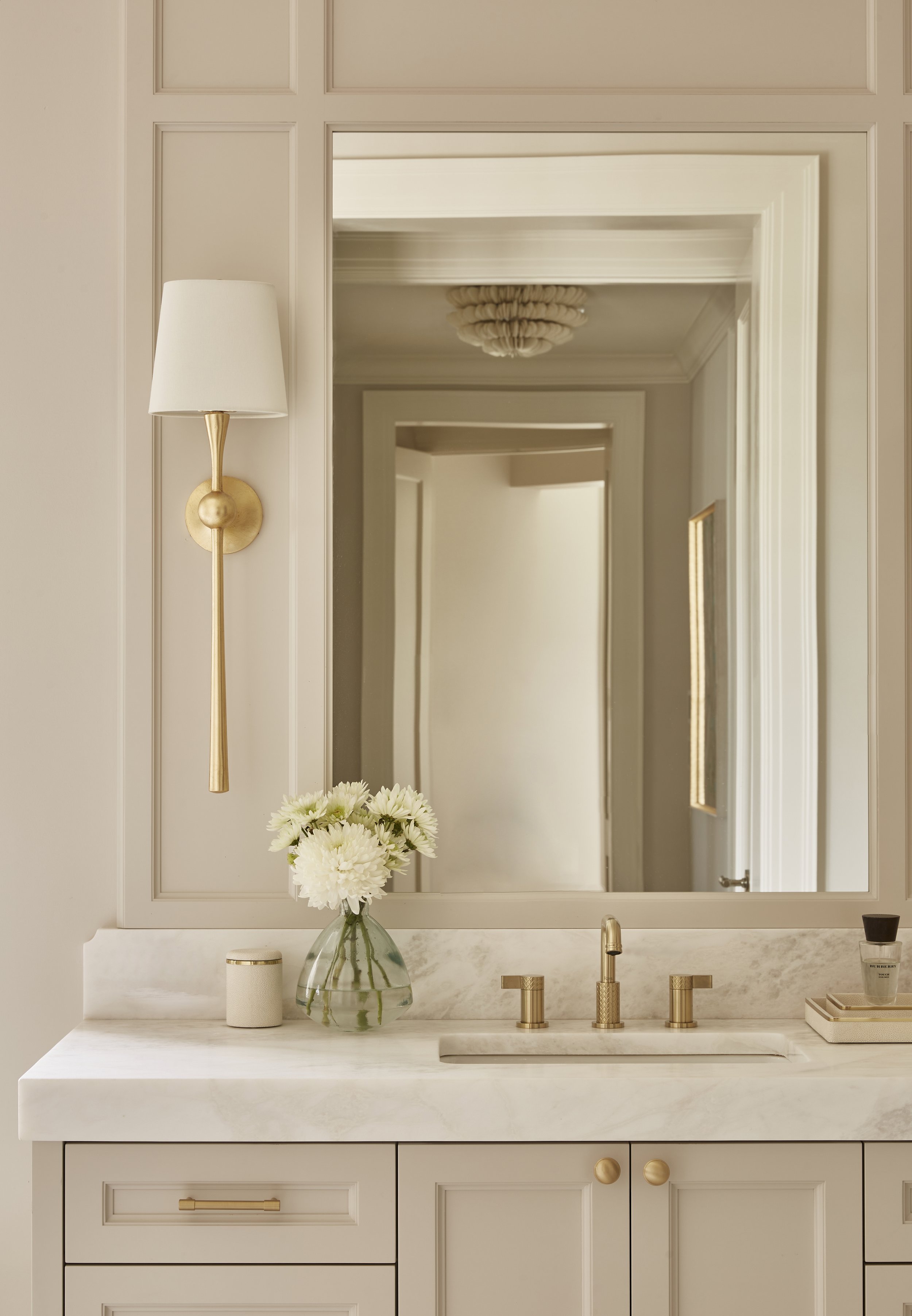 A bathroom vanity with a marble countertop, a large mirror, a vase of white flowers, and gold fixtures, illuminated by a wall sconce on the left.