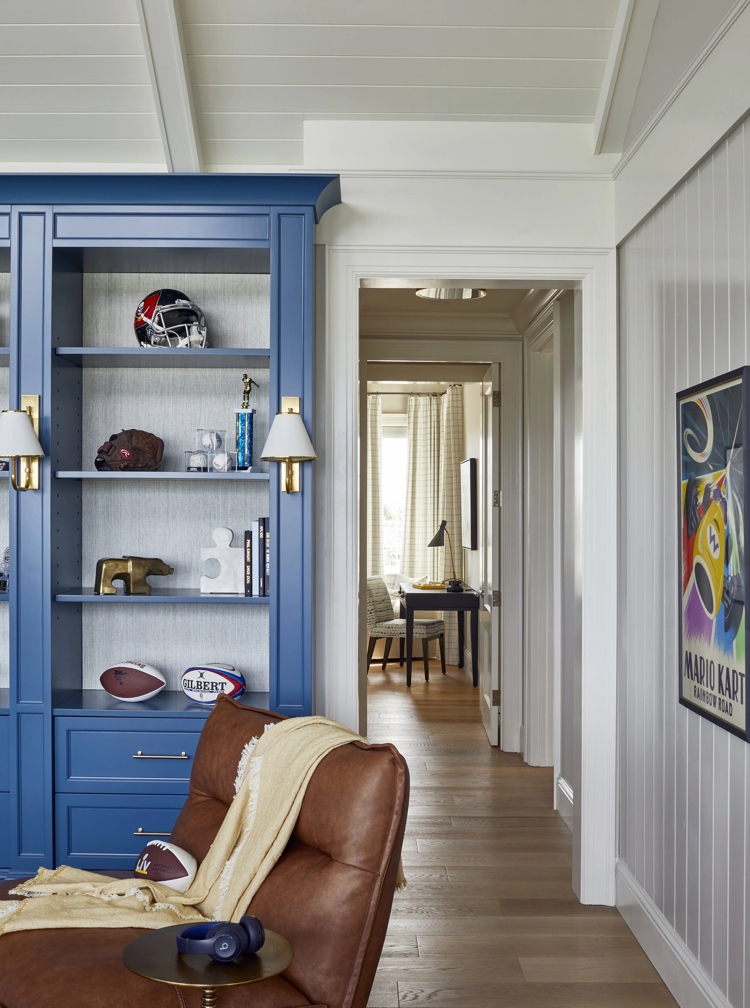 Living room with a blue bookshelf displaying football helmets and other sports memorabilia, a tan armchair with a blanket and footballs, and a hallway leading to a room with a desk and chair. A colorful Mario Kart poster decorates the wall.