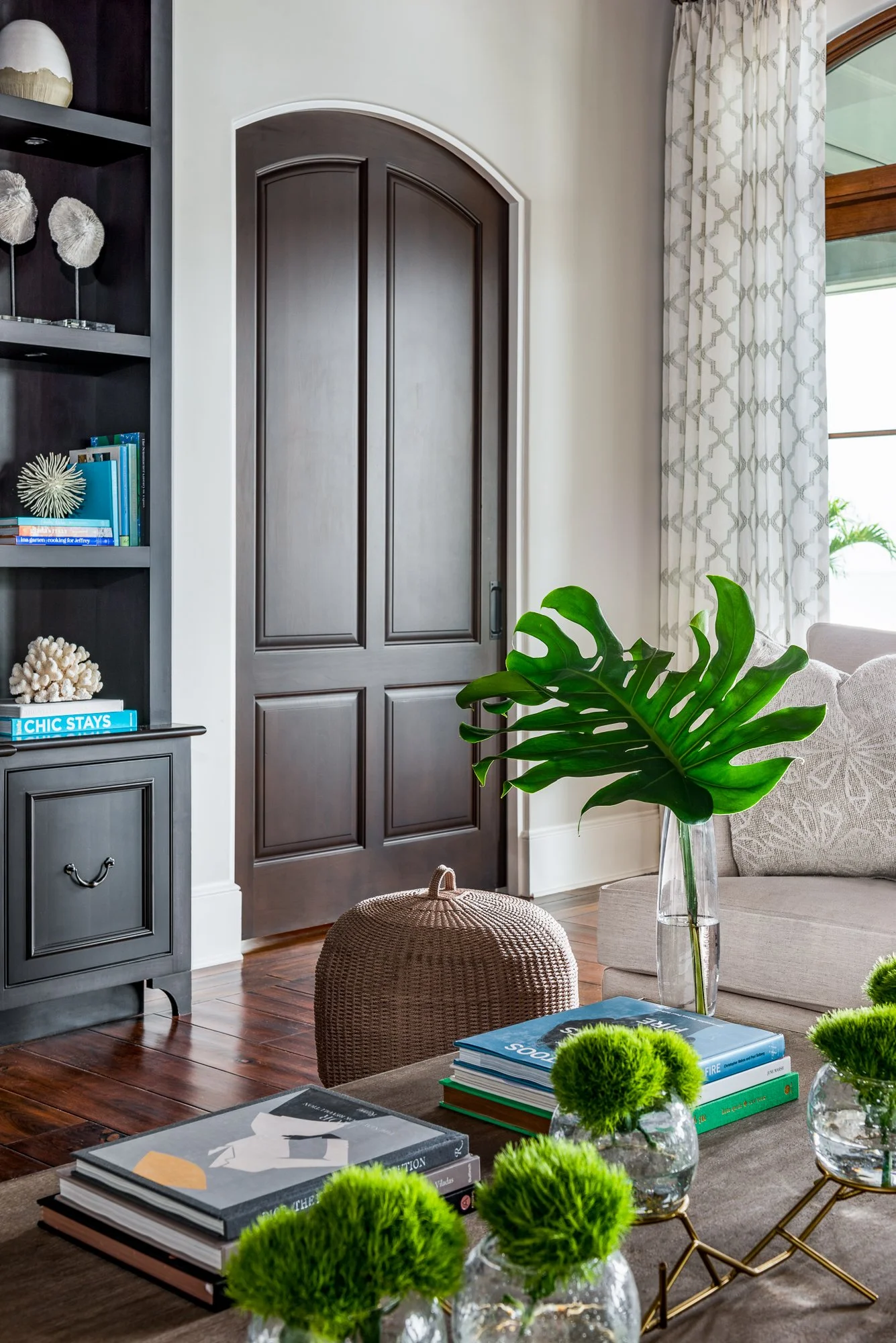 A living room corner with a closed dark wooden door, a beige sofa with patterned cushions, a glass vase with a large green monstera leaf, a woven ottoman, a brown wooden table with books, and small glass vases with green moss balls, illuminated by na