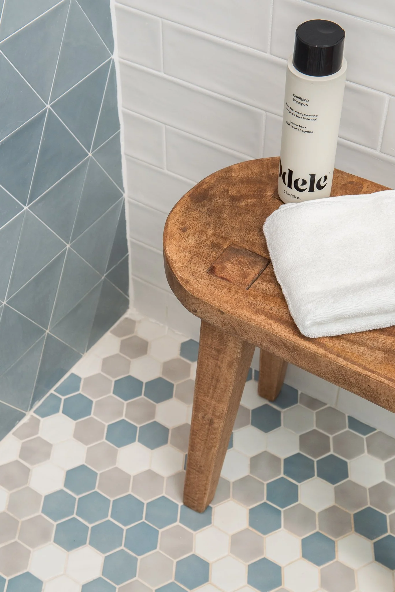 A wooden stool with a white towel and a bottle of clarifying shampoo on top, set against a tiled bathroom wall and floor.
