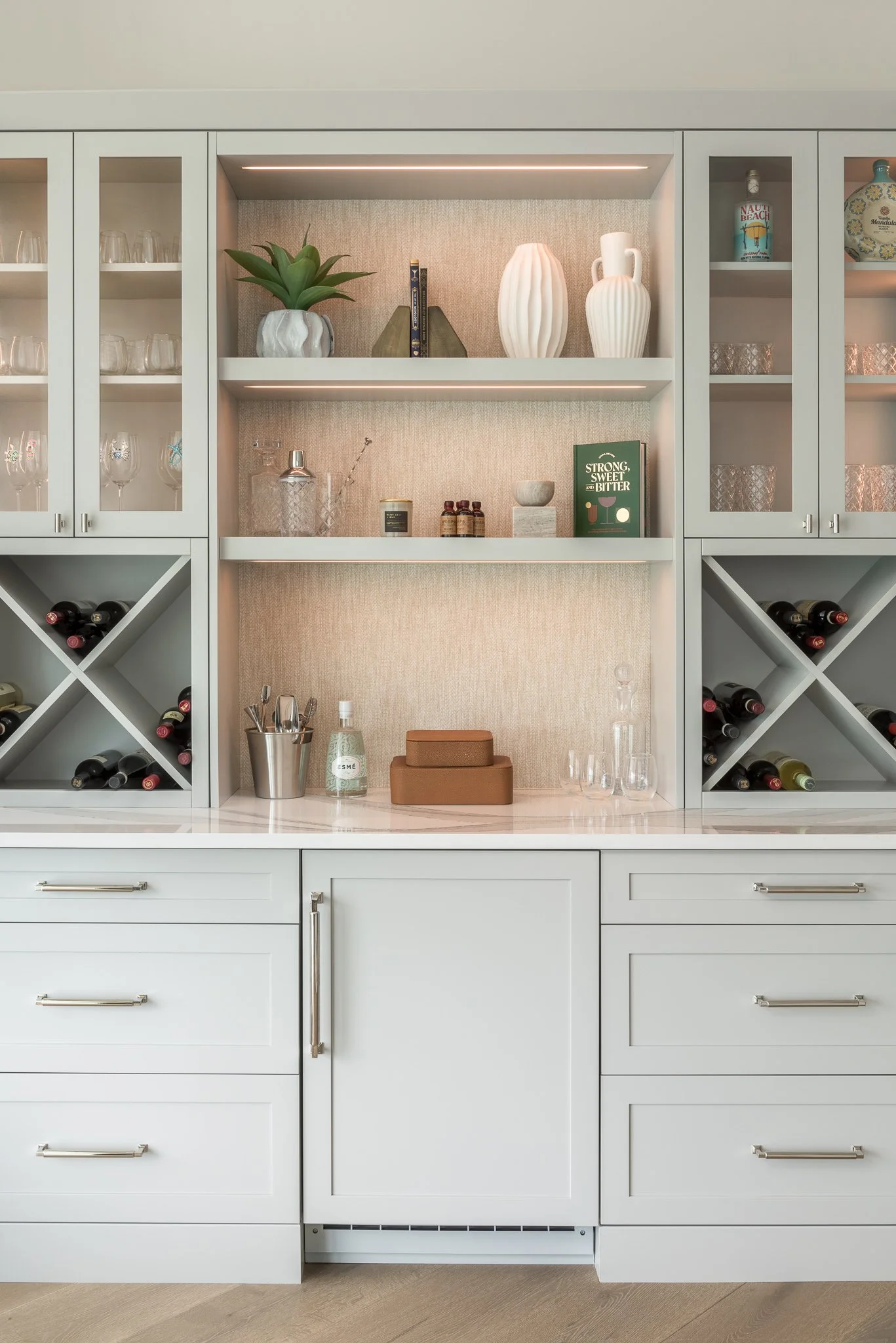 White kitchen cabinet with glass doors, wine racks, and decorative vases, glasses, and books displayed on open shelves.