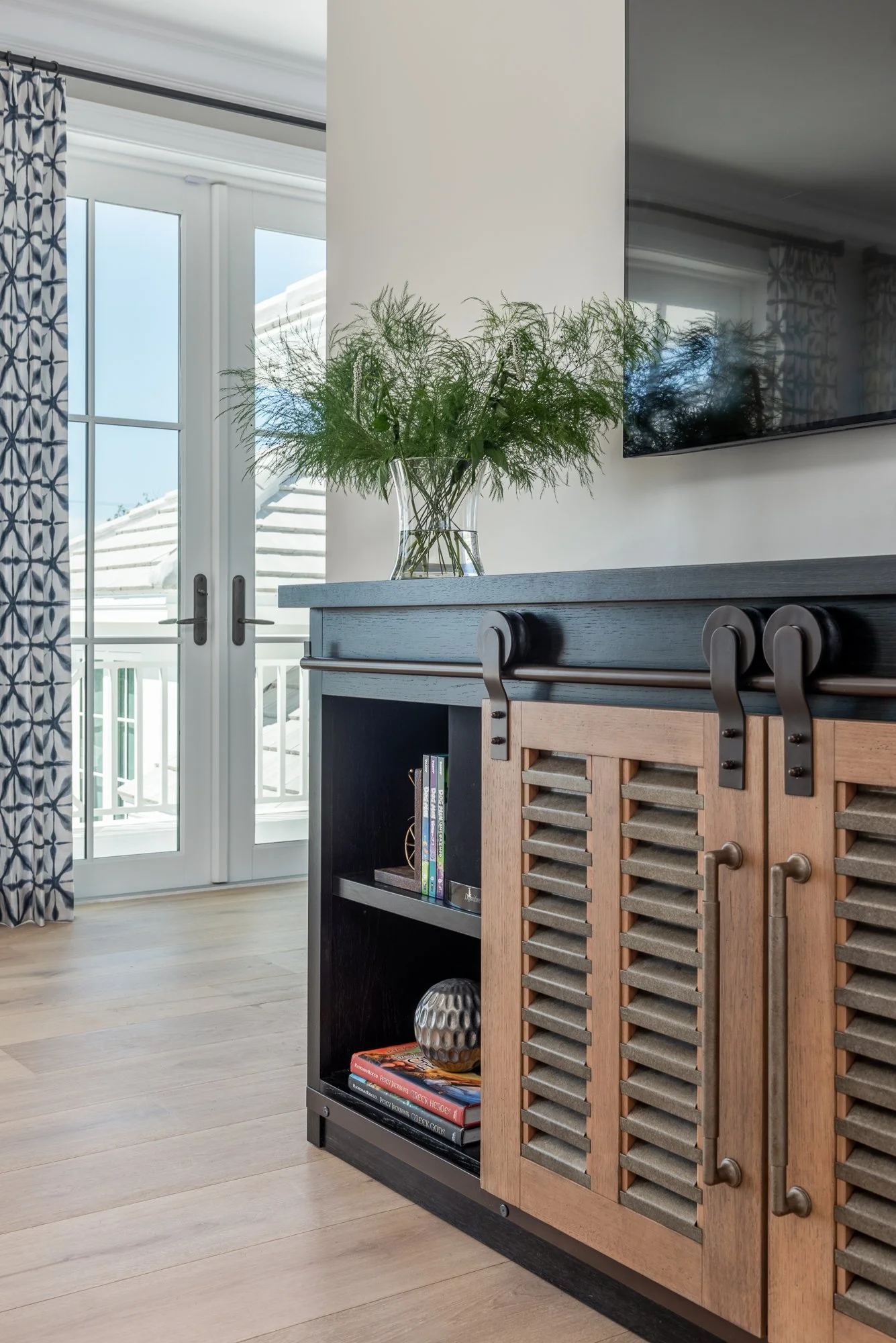 Interior of a living room with a sliding glass door, patterned curtains, a black and wood cabinet, a green plant, and a wall-mounted television.