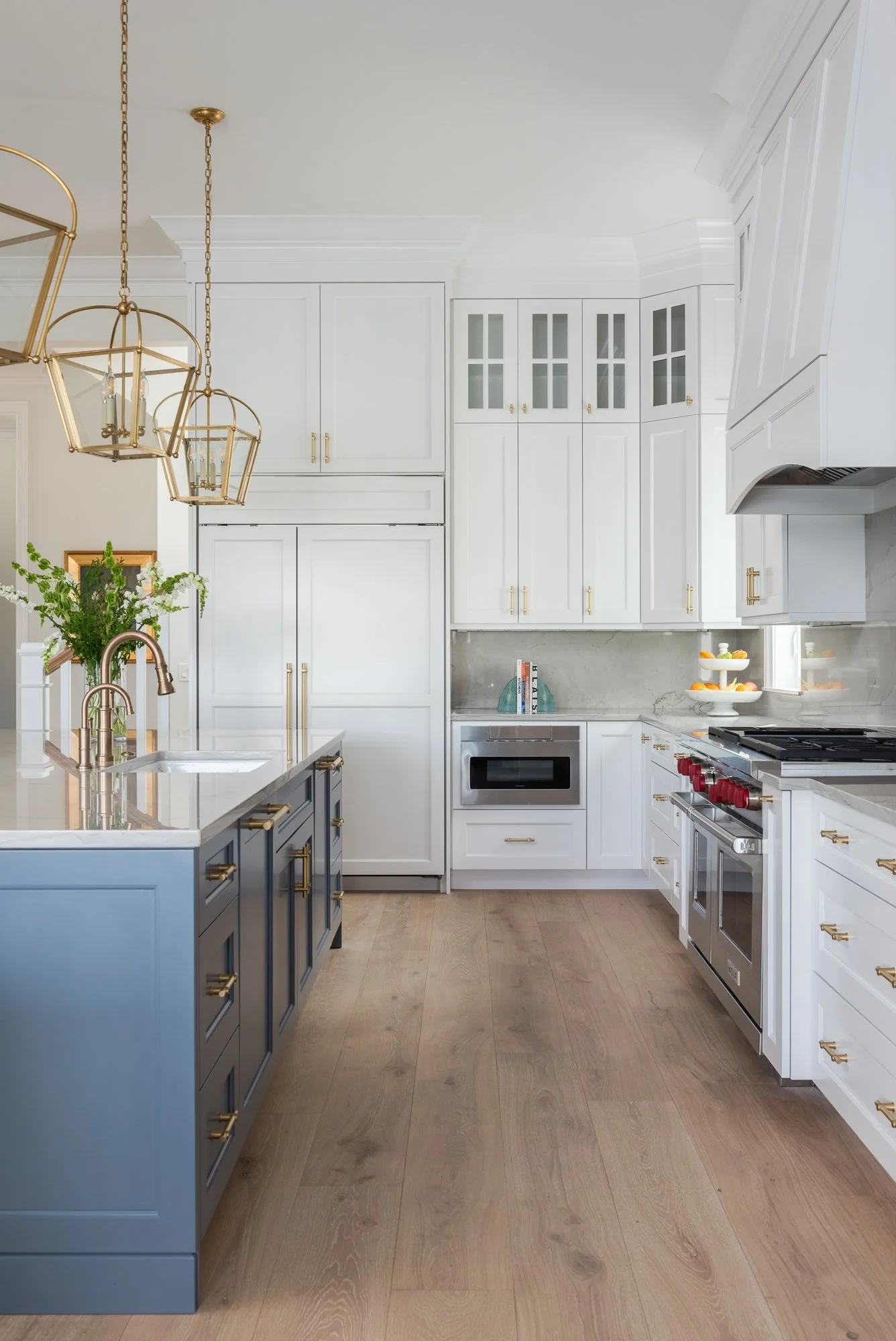 A bright, modern kitchen with white cabinets, gold hardware, a gray island with a gold sink faucet, and a black stove with red knobs. There are hanging gold pendant lights and a small window near the stove. A microwave and some books are on the count