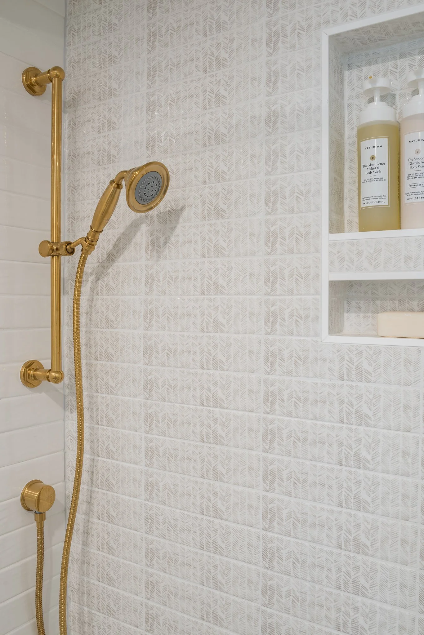 Shower with a gold-colored shower head and handheld spray, built-in shelf with body wash and soap, tiled wall with light beige and white patterns.