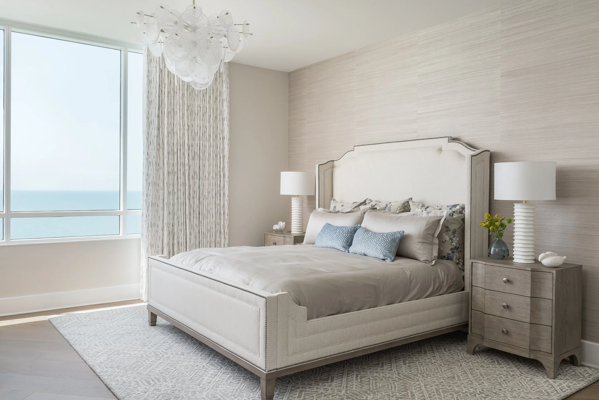 A modern bedroom with a large bed, beige bedding, blue decorative pillows, two bedside tables with lamps, plants, and shells, light-colored curtains, and a large window with ocean view.