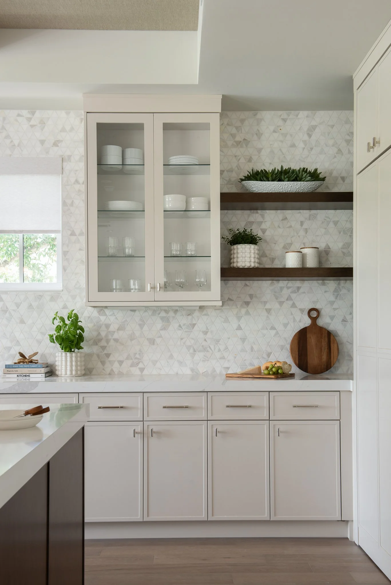 Kitchen with white cabinets, patterned backsplash, open shelves with white and clear dishes, and kitchen countertop with cutting board, plant, and books.