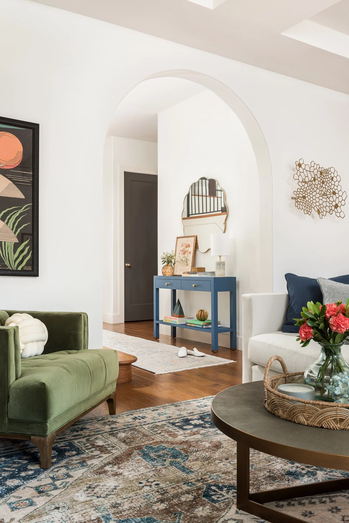 Living room with green velvet sofa, white couch with navy and gray pillows, round coffee table with a vase of pink flowers, framed wall art, blue console table with decorative items, mirror and wall art, hardwood floors, and patterned rug.