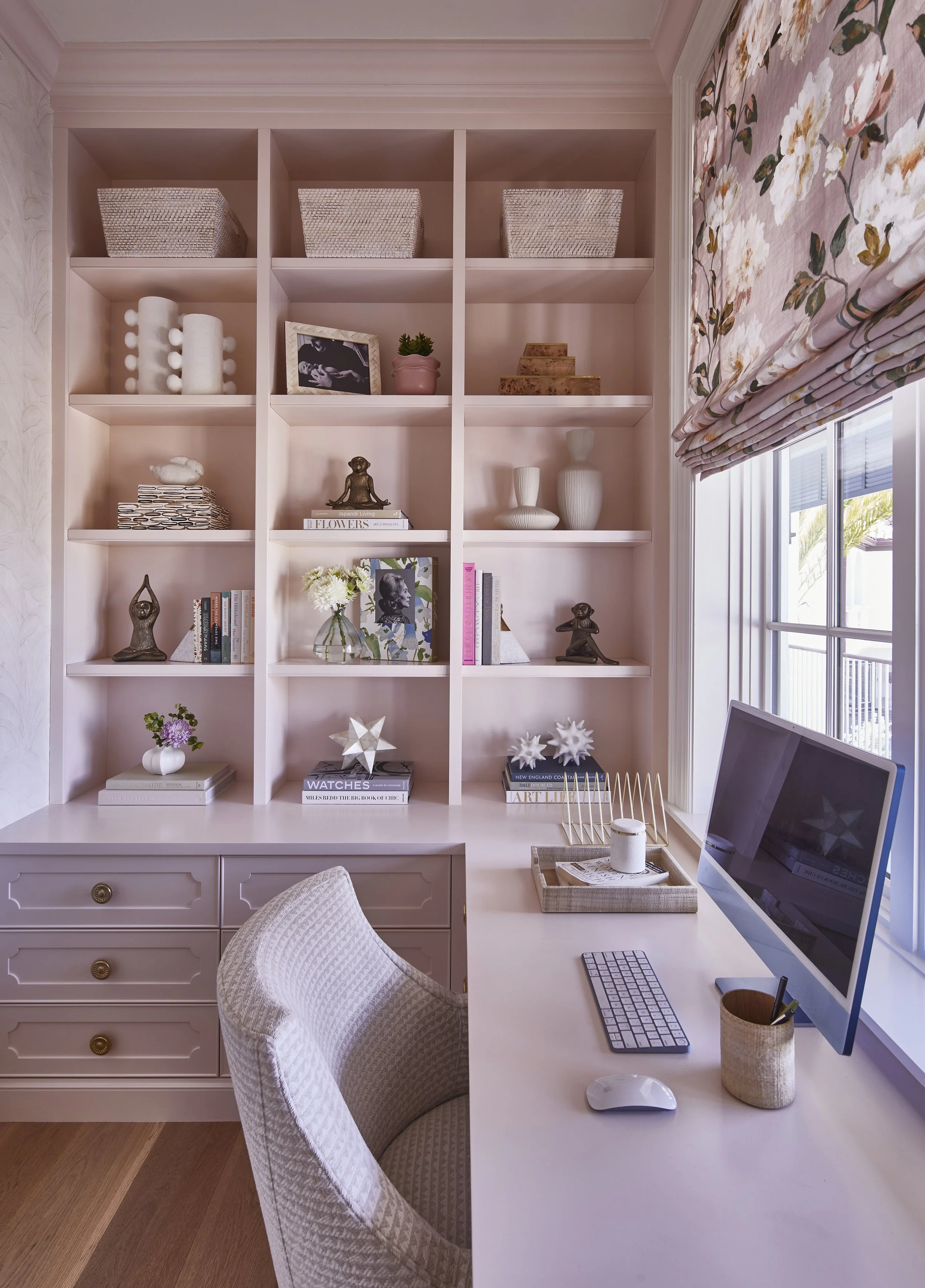 Home office workspace with built-in pink shelving filled with decorative items like vases, books, and art pieces. A white desk with a computer monitor, keyboard, mouse, and office supplies. A white upholstered chair and a window with floral Roman sha