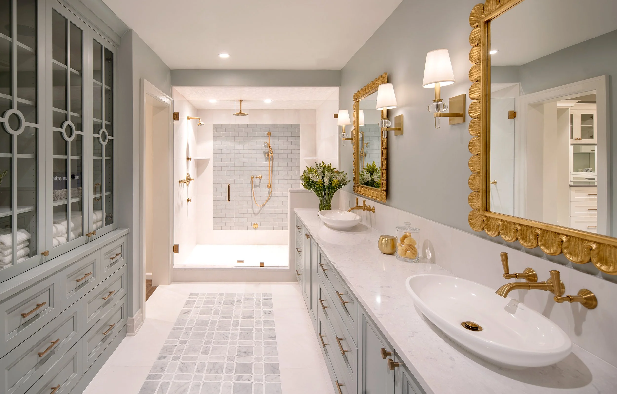 A luxurious bathroom with light gray walls, a double sink vanity with a white marble countertop, two gold-framed mirrors, and wall-mounted gold lights. There are two oval vessel sinks, a vase with flowers, and decorative jars. The walk-in shower has 