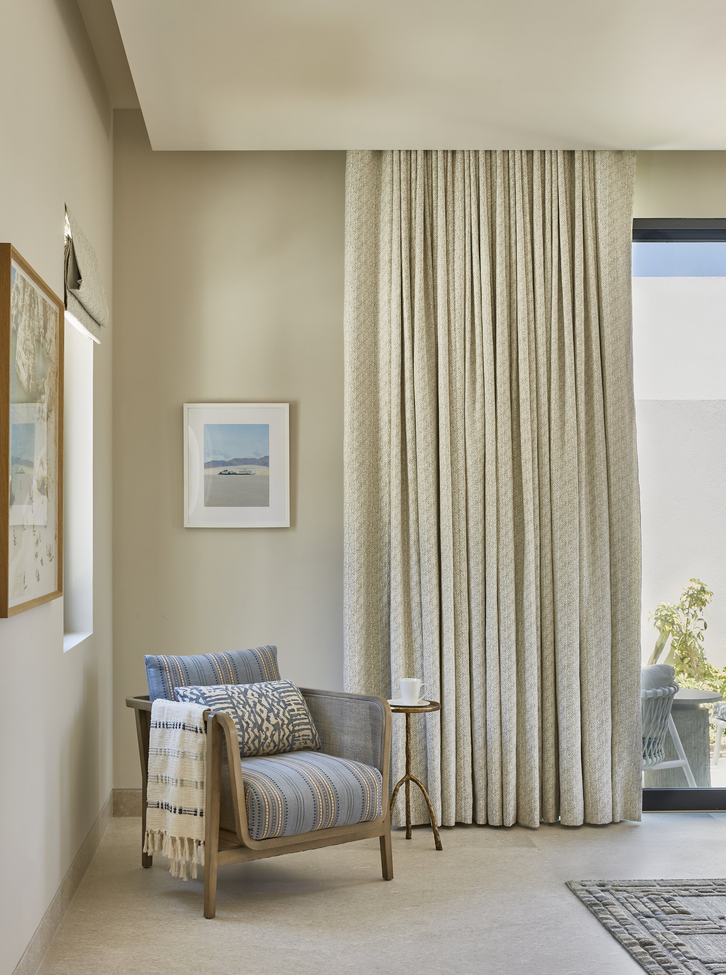 Living room corner with beige curtains, a small wooden side table, a patterned armchair with decorative pillows and a throw, and framed artwork on the wall.