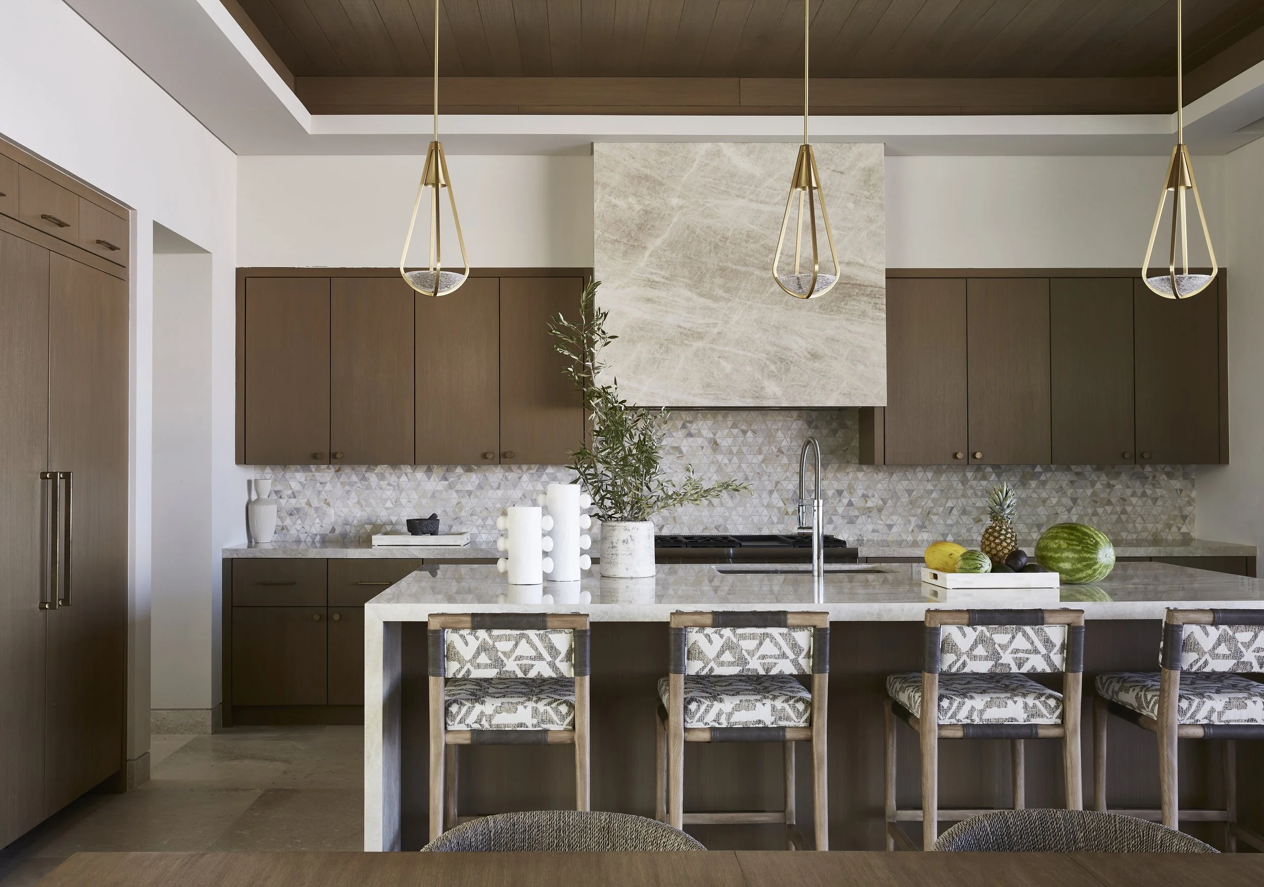 Modern kitchen with wooden cabinets, a marble island, and pendant lighting, decorated with vases and fresh fruit.