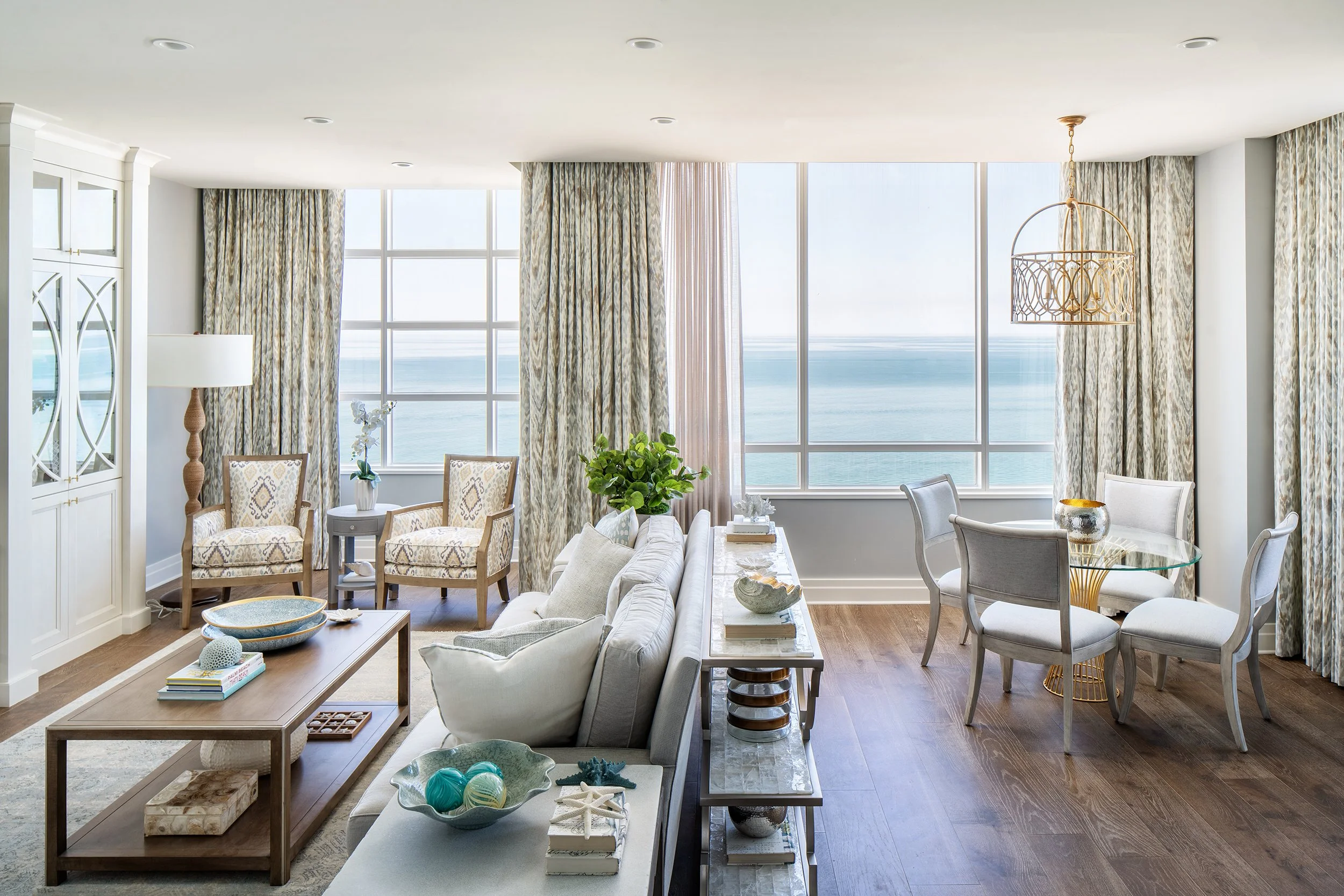 Living room with large windows overlooking the ocean, light-colored furniture, a round glass dining table with four chairs, and elegant decor.